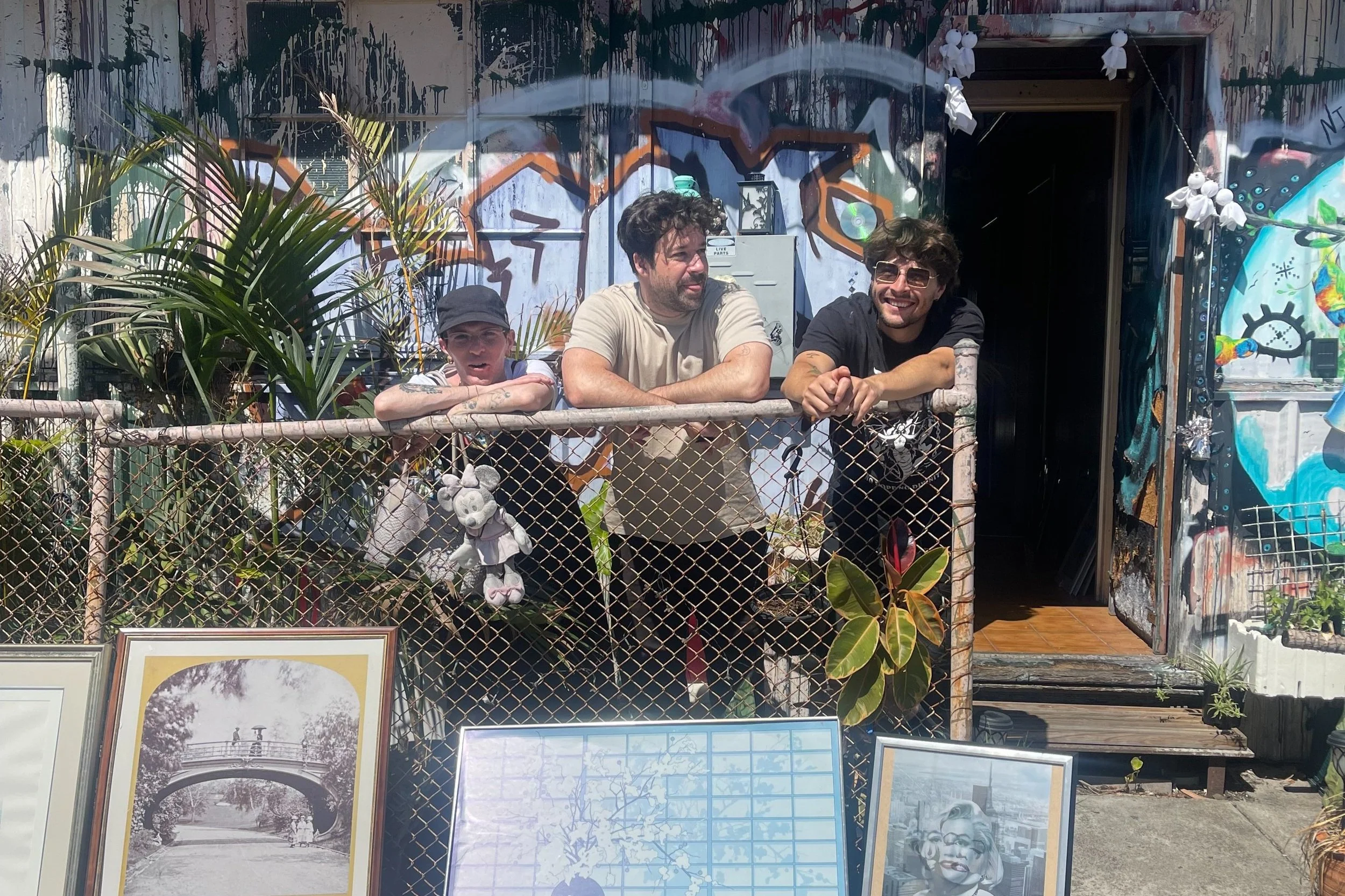 Three men leaning on a rusty fence with a graffitied wall behind them, surrounded by plants and framed photos at the front, outside a building.
