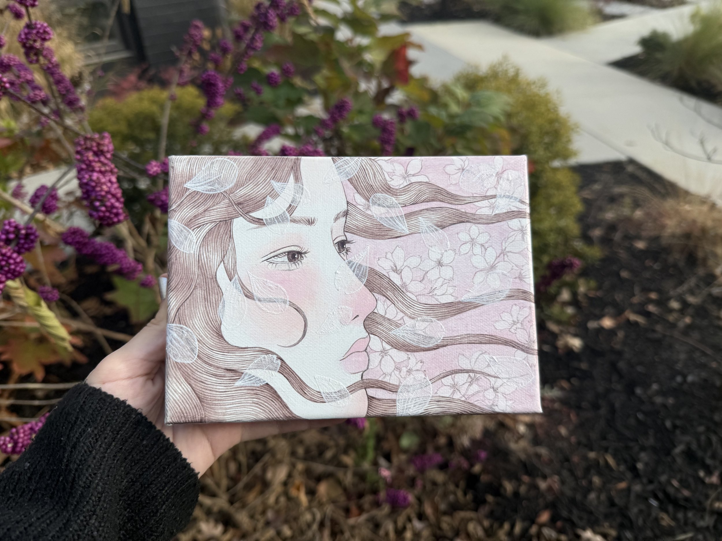 A person holding a decorative box featuring a line drawing of a woman's face with flowing hair, surrounded by flower and leaf patterns, outdoors with purple flowering bushes and a garden path in the background.