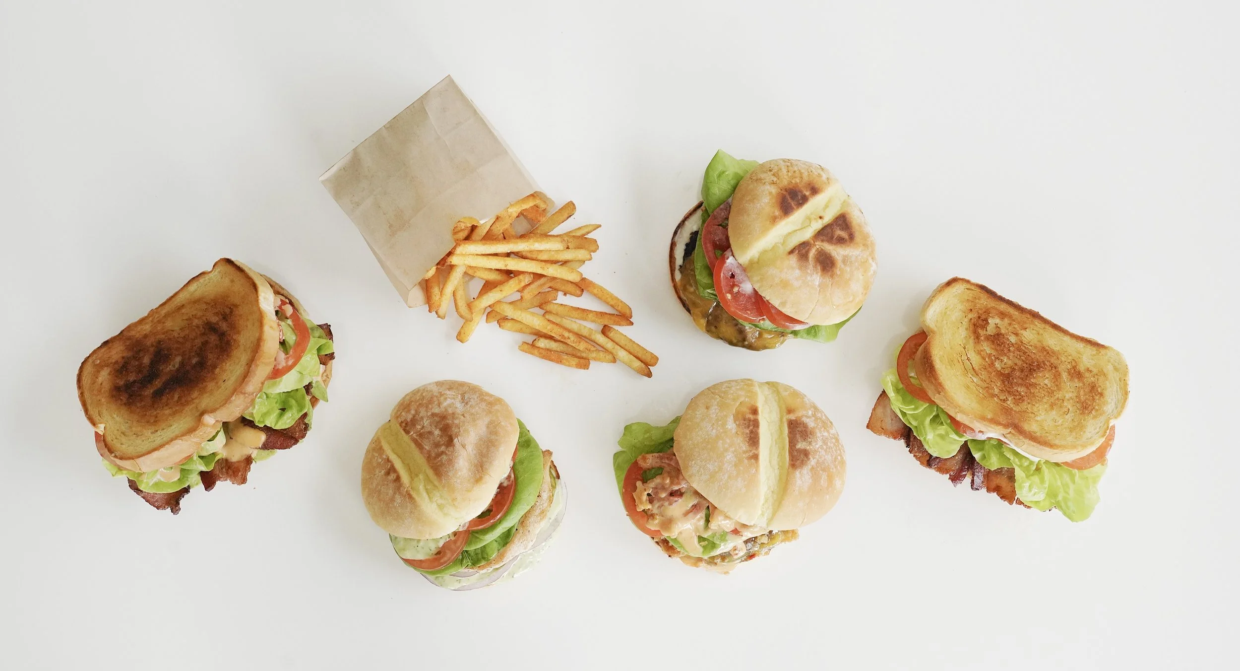 Several mini sandwiches with lettuce, tomato, cheese, and meats, along with a paper cone of French fries on a white surface.