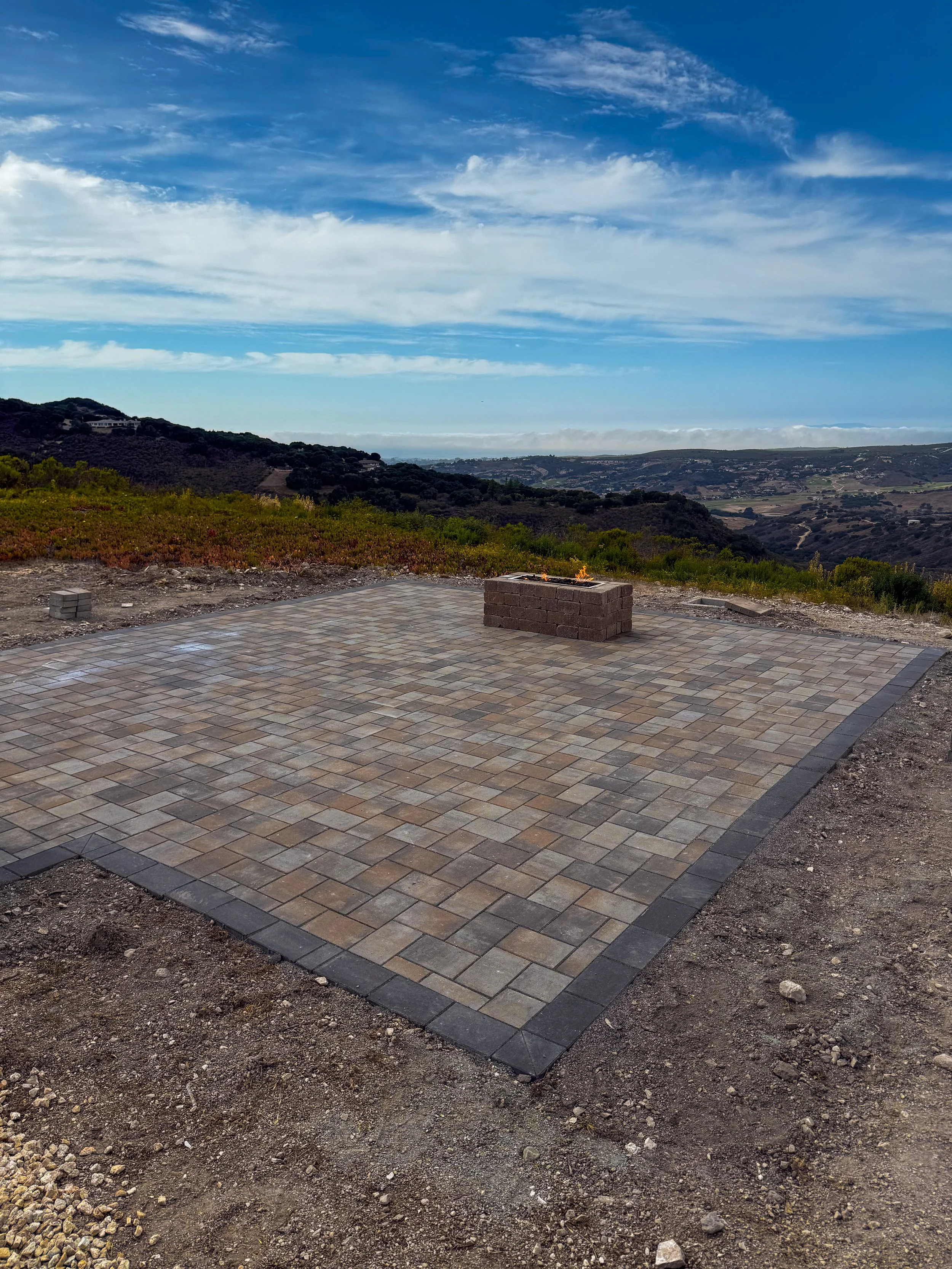 Patio with interlocking bricks and a fire pit in the foreground, overlooking a landscape of rolling hills and a cloudy sky.