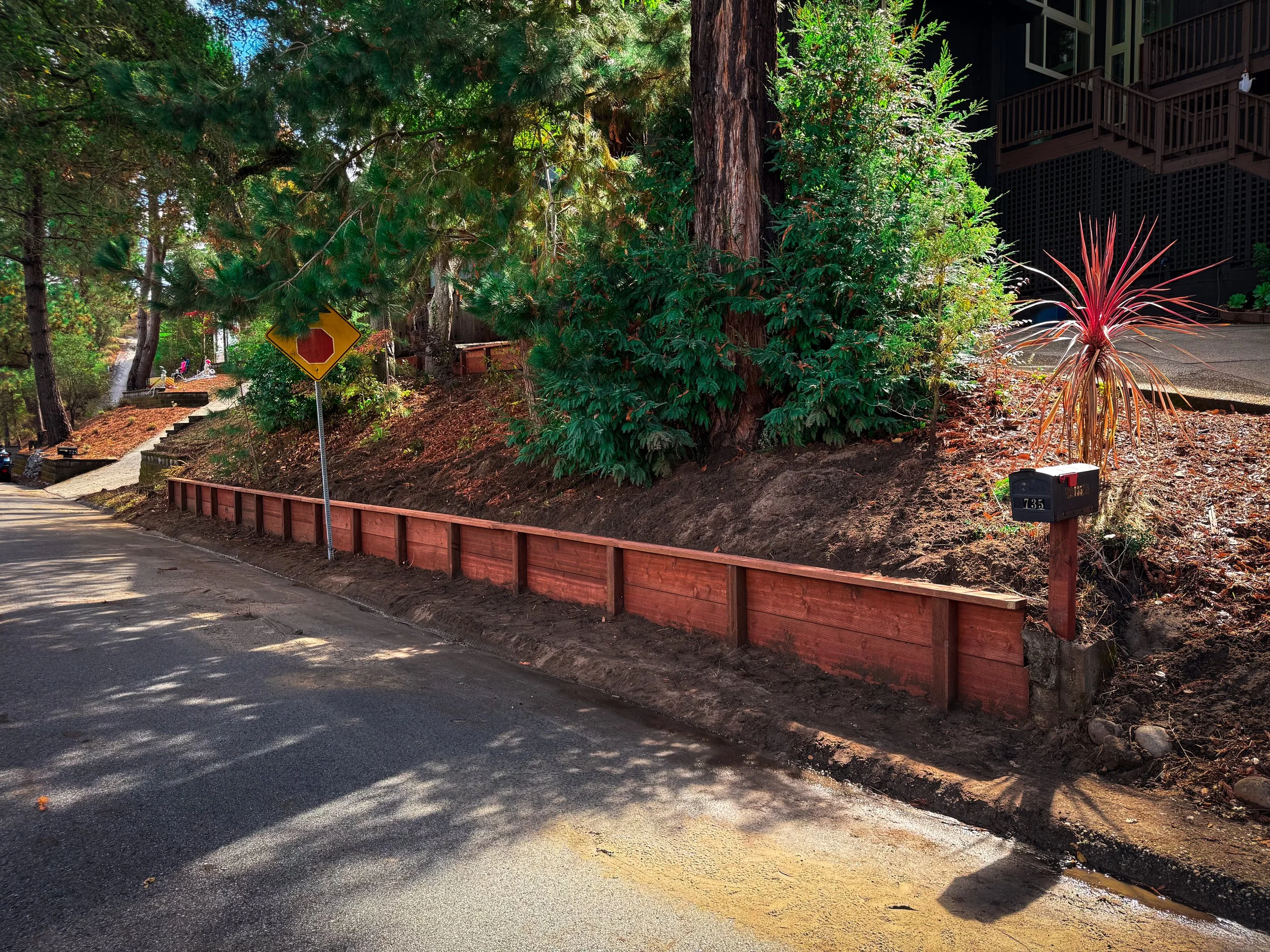 A sidewalk with a red wooden fence on the right side, tall green trees and bushes, a stop sign partially covered by foliage, and a dark residential building in the background.