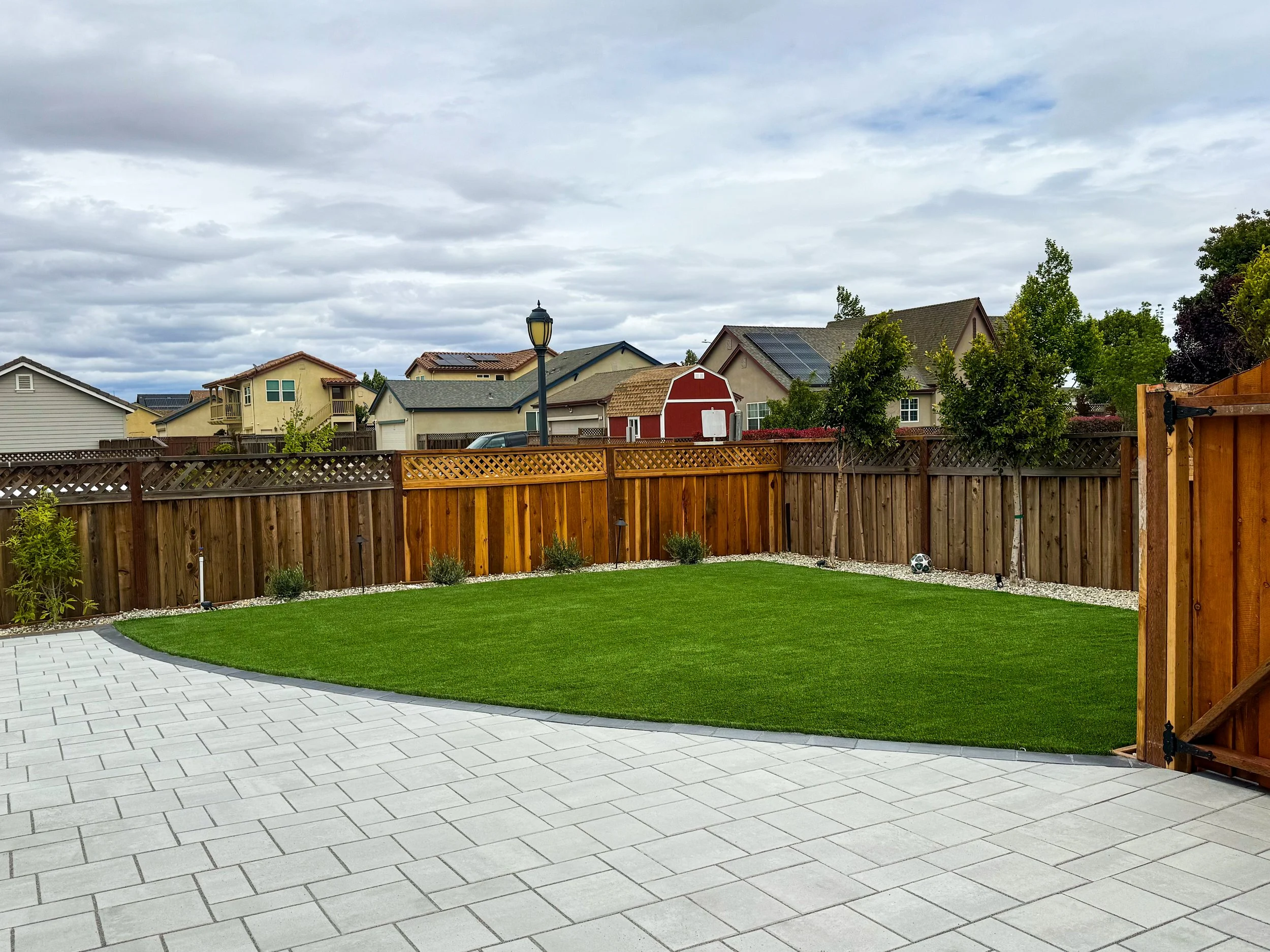 A backyard with a brick patio, green lawn, wooden fence, trees, and a cloudy sky.