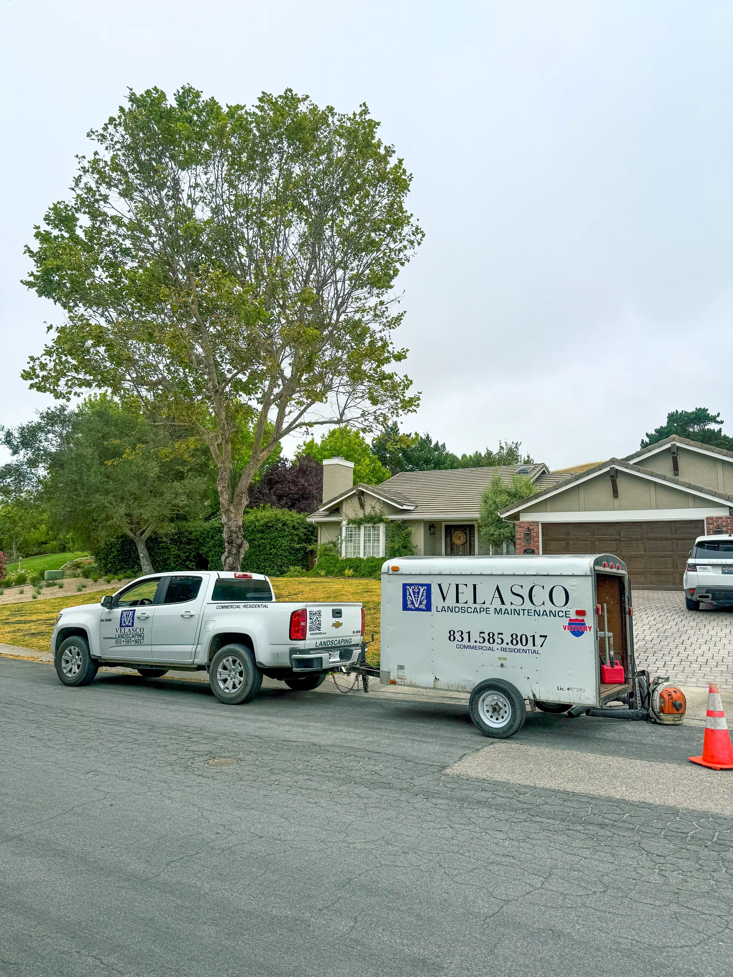 A white pickup truck and a white trailer with landscaping company branding parked on a residential street. The trailer is connected to the truck, and there is an orange traffic cone on the street near the trailer. A house with a sloped roof, a chimney, and a garage is visible in the background, along with a large tree and other greenery.