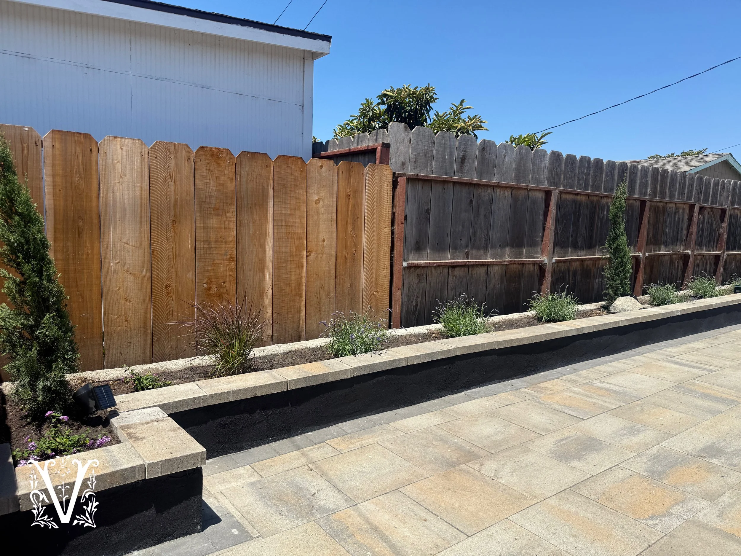 Wooden and wire fence with plants and small trees along the sidewalk in front of a white building and a clear blue sky.