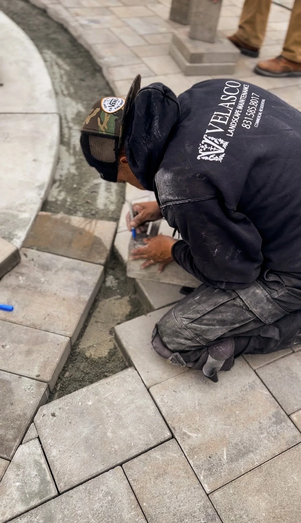 Construction worker installing paving stones on a sidewalk, kneeling and using tools to set stones in place, wearing a black shirt and camouflage cap.