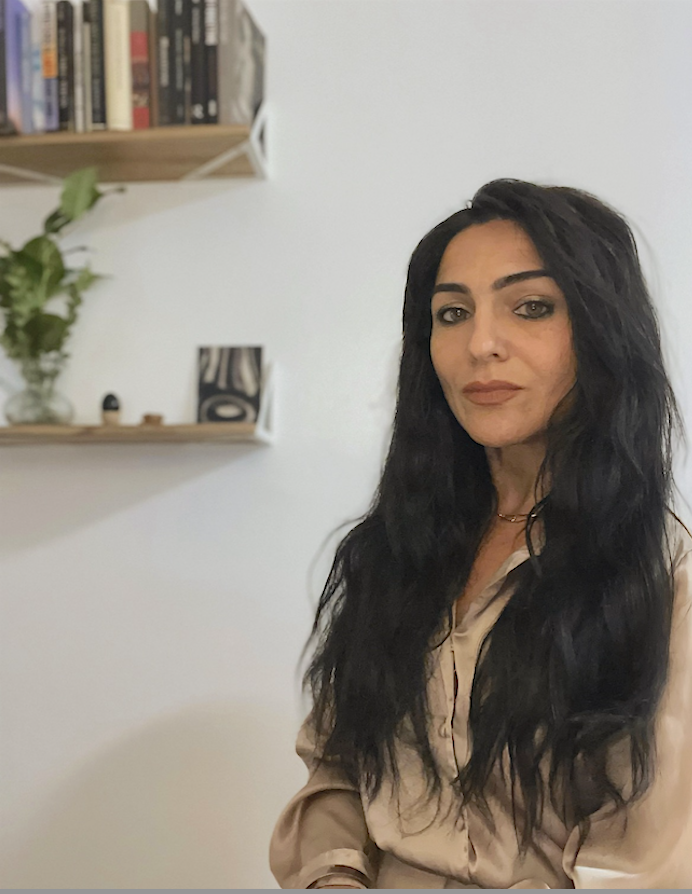 A woman with long, wavy black hair and light brown skin, wearing a beige blouse, sits indoors with a white wall and a wooden shelf with books and a plant in the background.