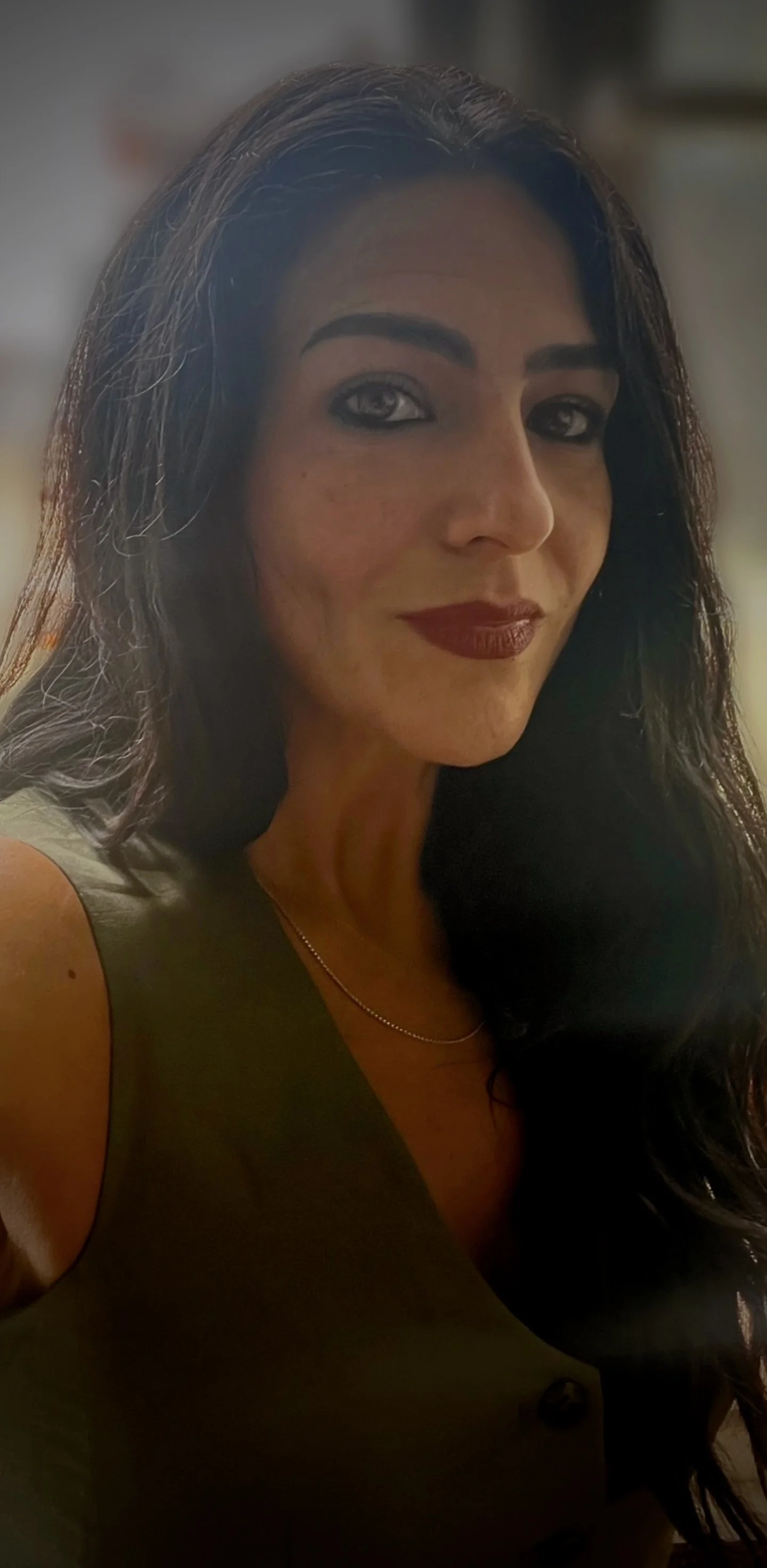 A woman with long dark hair and makeup, wearing a sleeveless top and a delicate necklace, looking at the camera with a slight smile.
