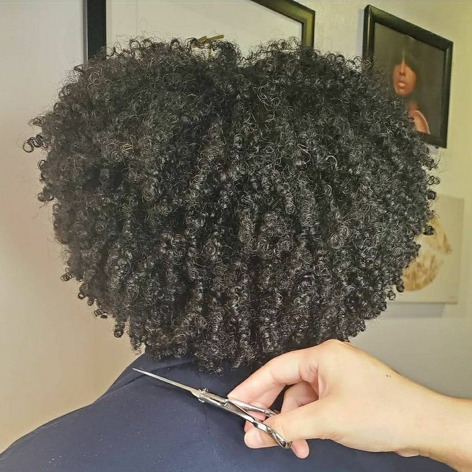 A person with short, curly hair getting a haircut, with a hand holding scissors.