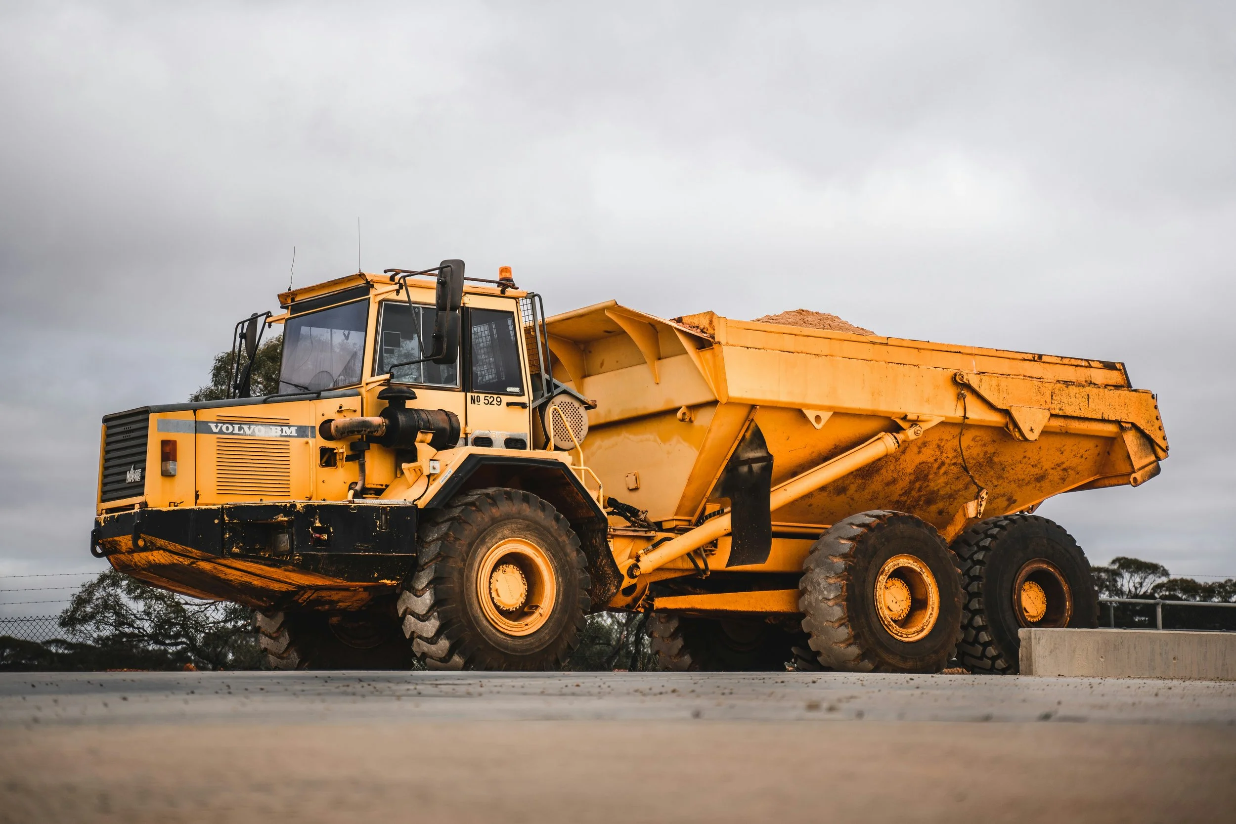A yellow Volvo dump truck parked on a construction site with a gray overcast sky in the background.