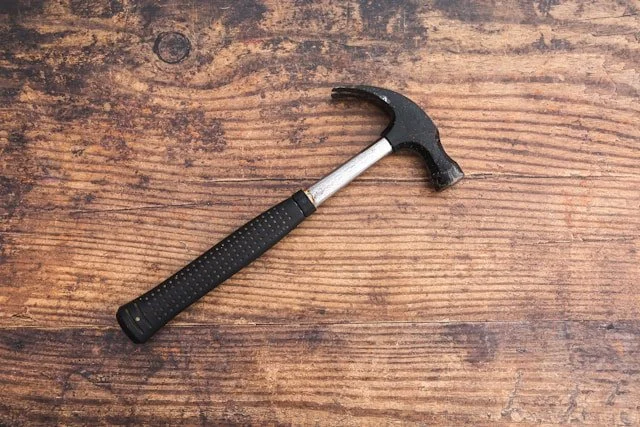 A small hammer with a black handle and a silver shaft, resting on a wooden surface.