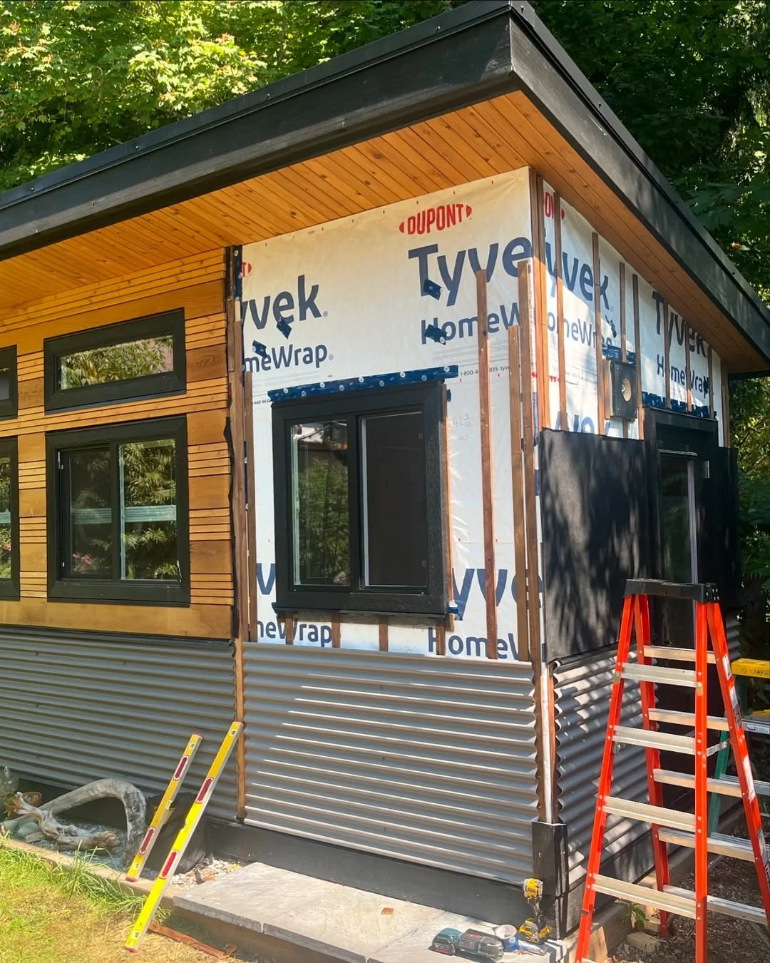 Partially constructed building with Tyvek sheathing, wooden siding, a flat roof, and several windows. A red ladder and other construction tools are visible nearby.