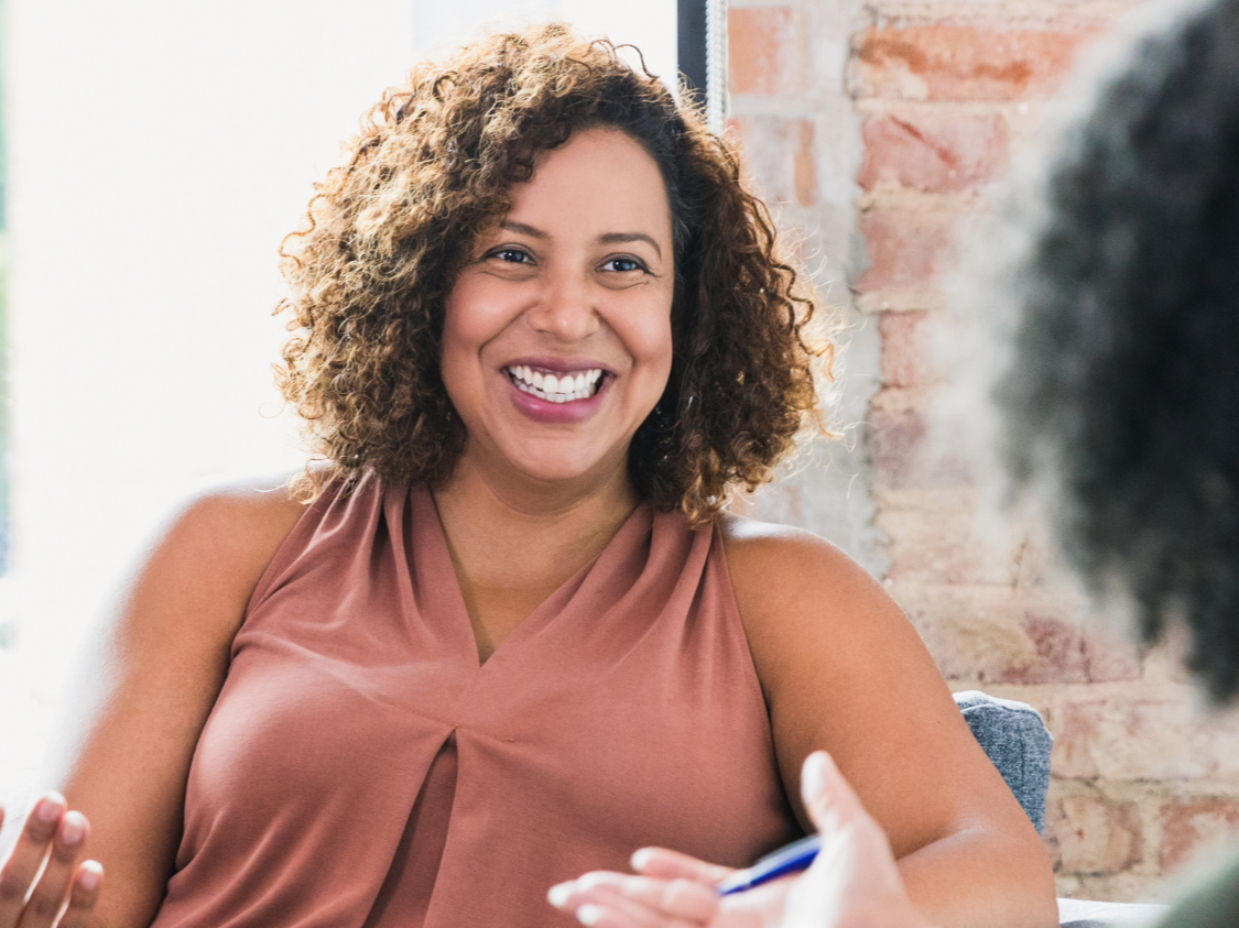 Smiling woman in a sleeveless brown top talking with another person in conversation.