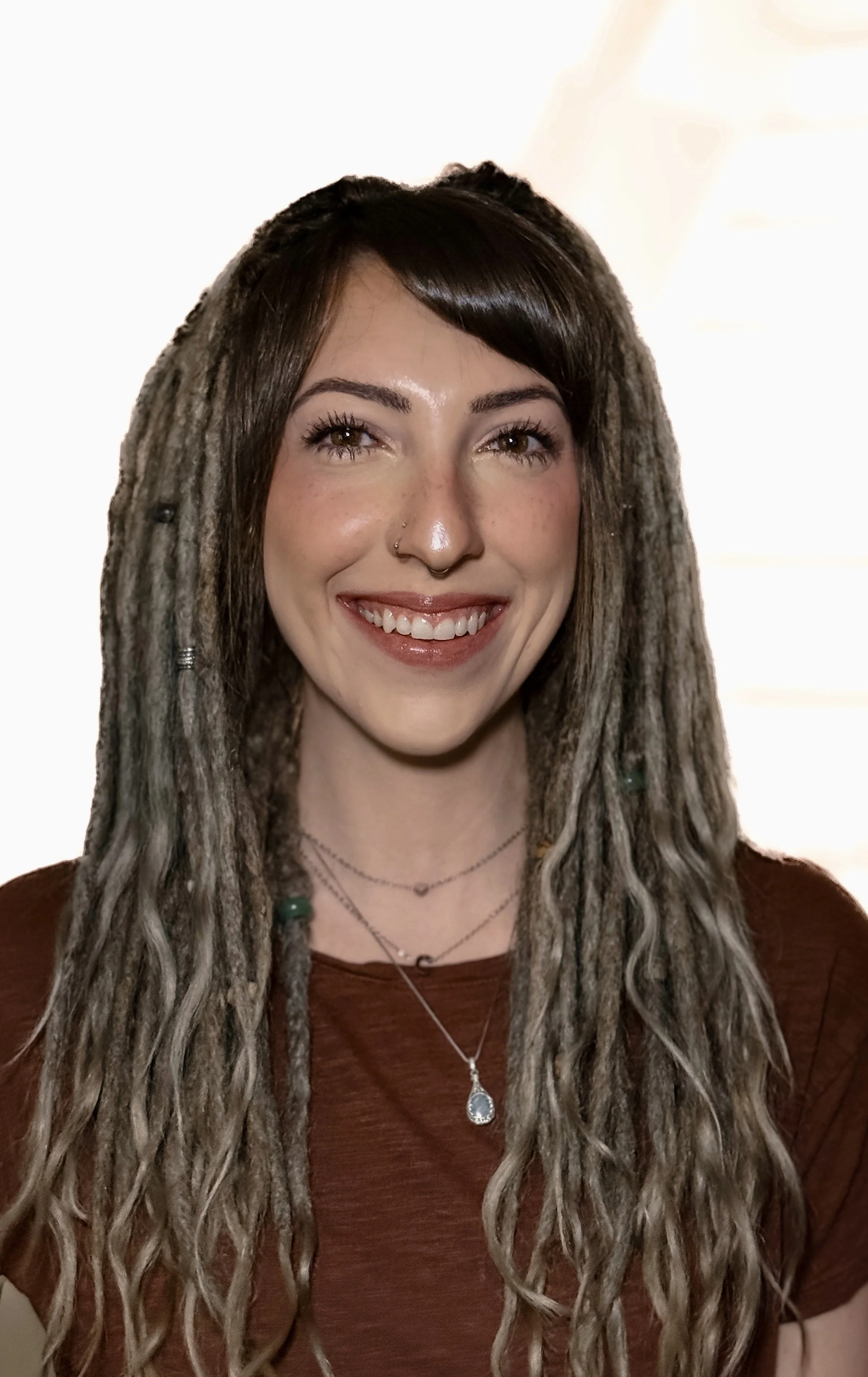A woman with long, gray, dreadlocked hair smiling at the camera, wearing a brown top and layered necklaces.