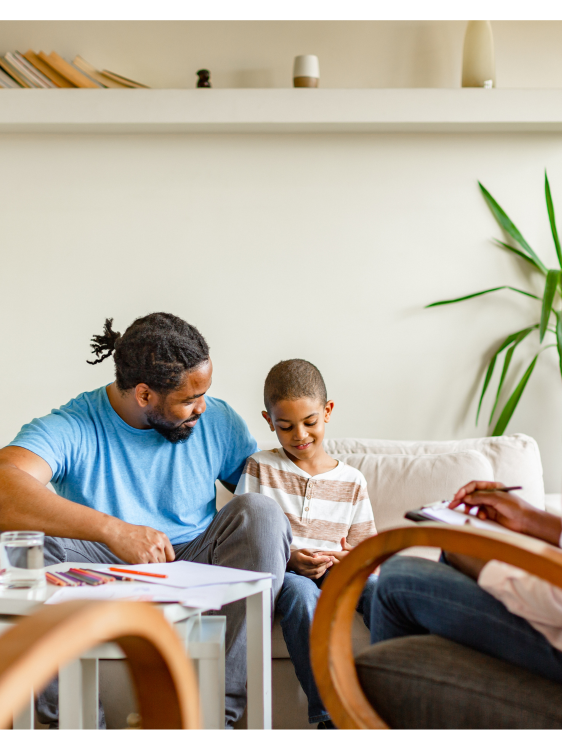 A therapy or counseling session with a family, including a therapist taking notes while a father and son sit on a sofa.