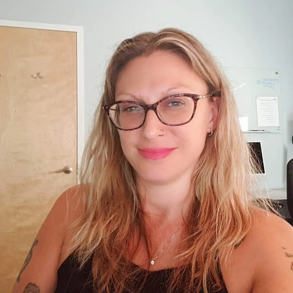 Close-up of a smiling woman with glasses, long wavy hair, and a nose piercing, in an office setting.