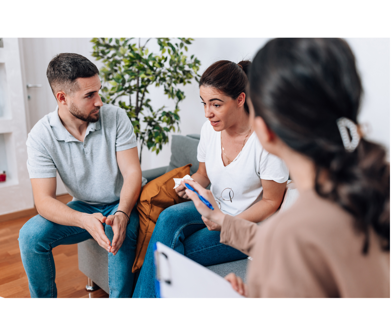 A woman in white shirt talking to a man in gray shirt during a counseling session on a gray sofa, with a counselor holding a clipboard and pen in front of her, in a living room setting with a green plant in the background.