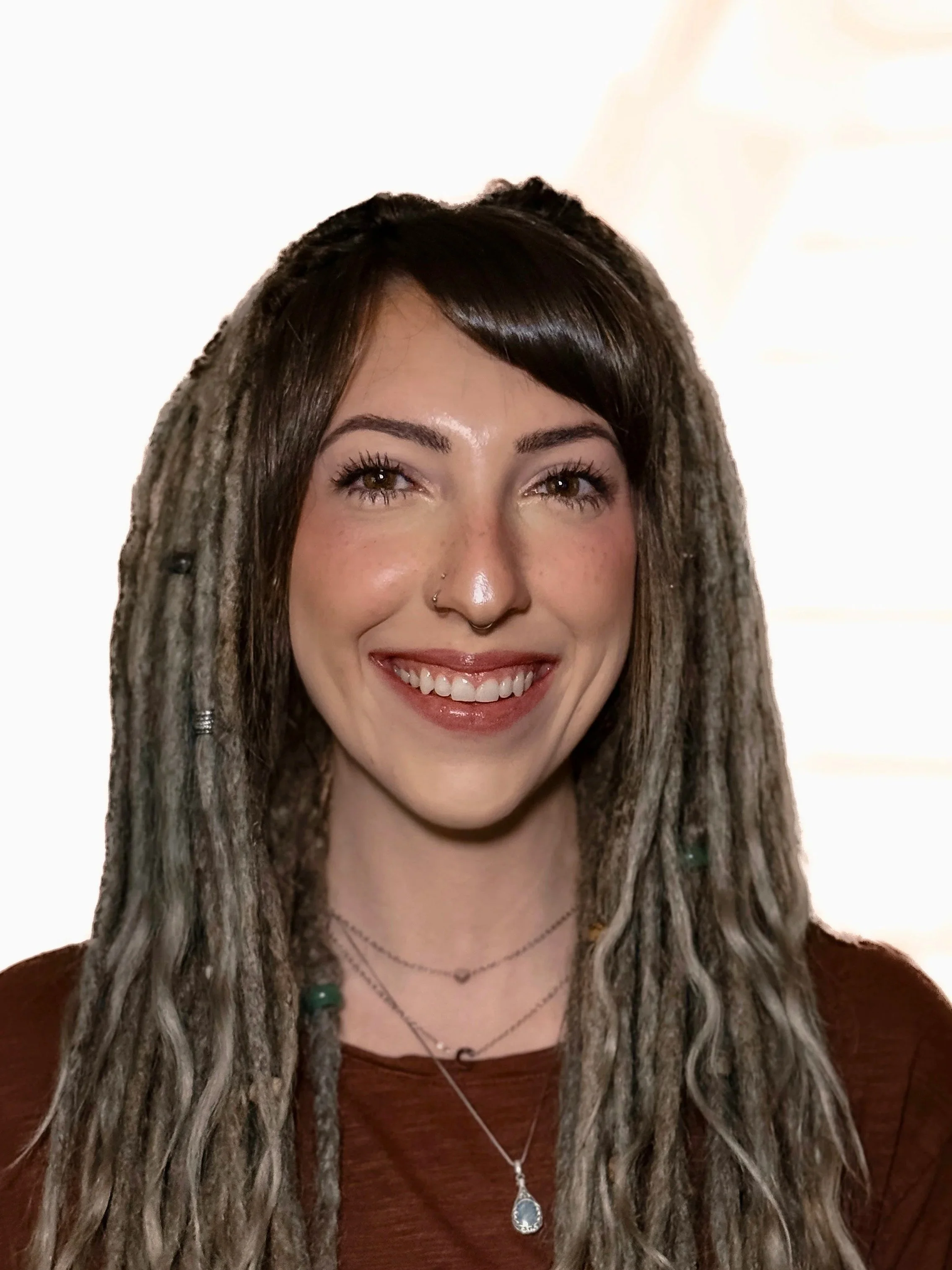 A smiling woman with dark brown hair styled in dreadlocks, wearing a nose ring, layered necklaces, and a brown top, standing in front of a bright, neutral background.