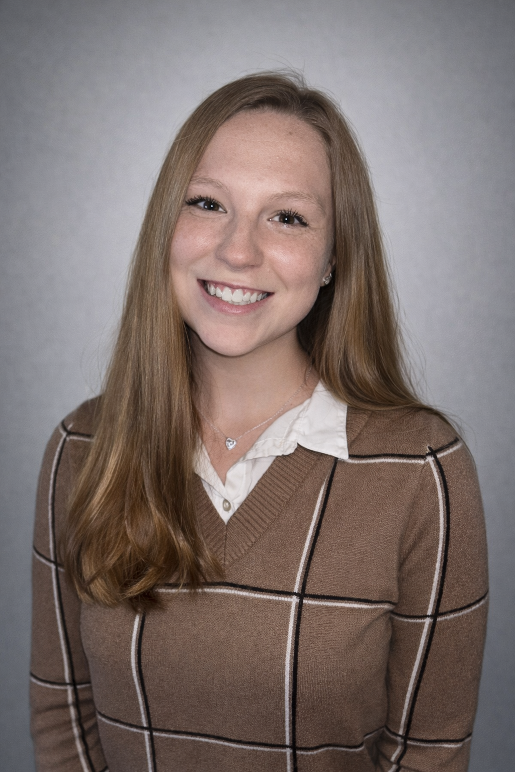 Portrait of a young woman with long red hair, smiling, wearing a brown plaid sweater over a white collared shirt and a heart-shaped necklace, set against a gray background.