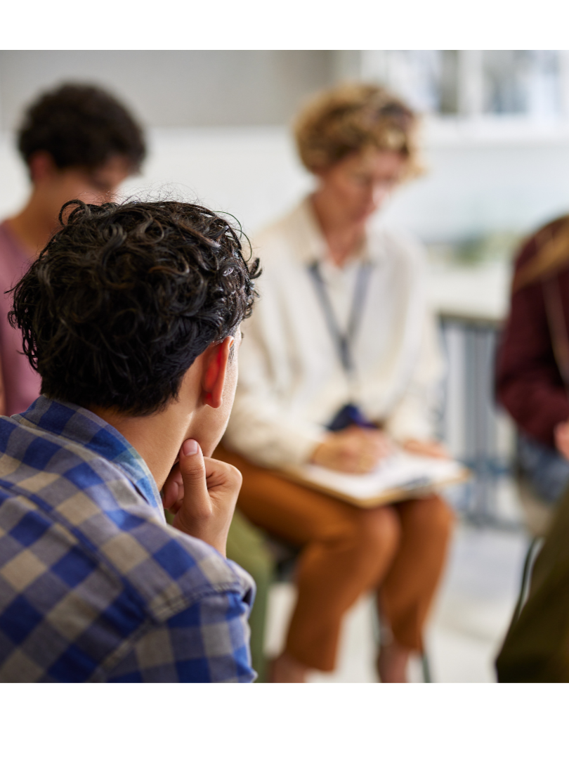 A group of people in a discussion, with a focus on a young man in a blue plaid shirt listening attentively.