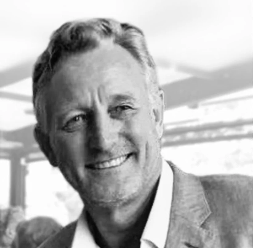 A black and white photo of a smiling middle-aged man with short hair, wearing a suit jacket and white shirt, in an indoor setting with blurred background.