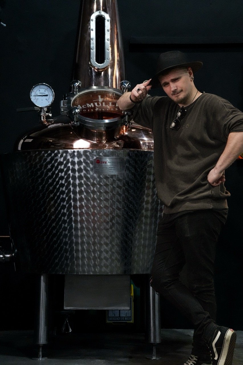 Man posing next to a large metal still, with gauges and pipes in a dimly lit room. The person is wearing a hat and casual clothing.