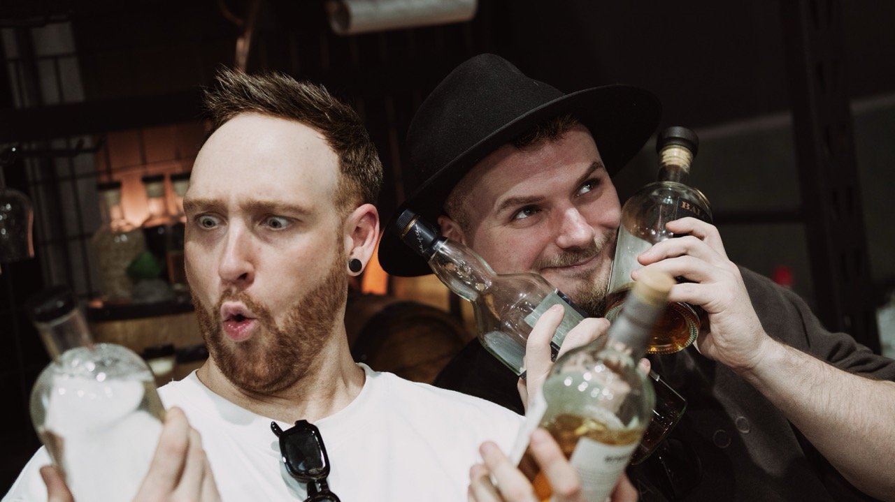Two men holding bottles of whisky with humorous expressions, one man wearing a black hat.