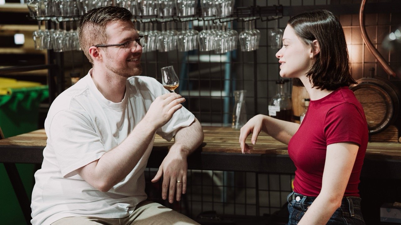 Two people sitting at a bar while one holds a glass of whiskey, surrounded by hanging glassware.