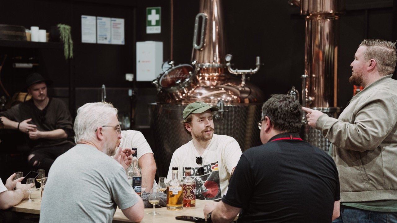 Group of people sitting around a table in a distillery, with bottles of liquor and a distillation apparatus in the background.