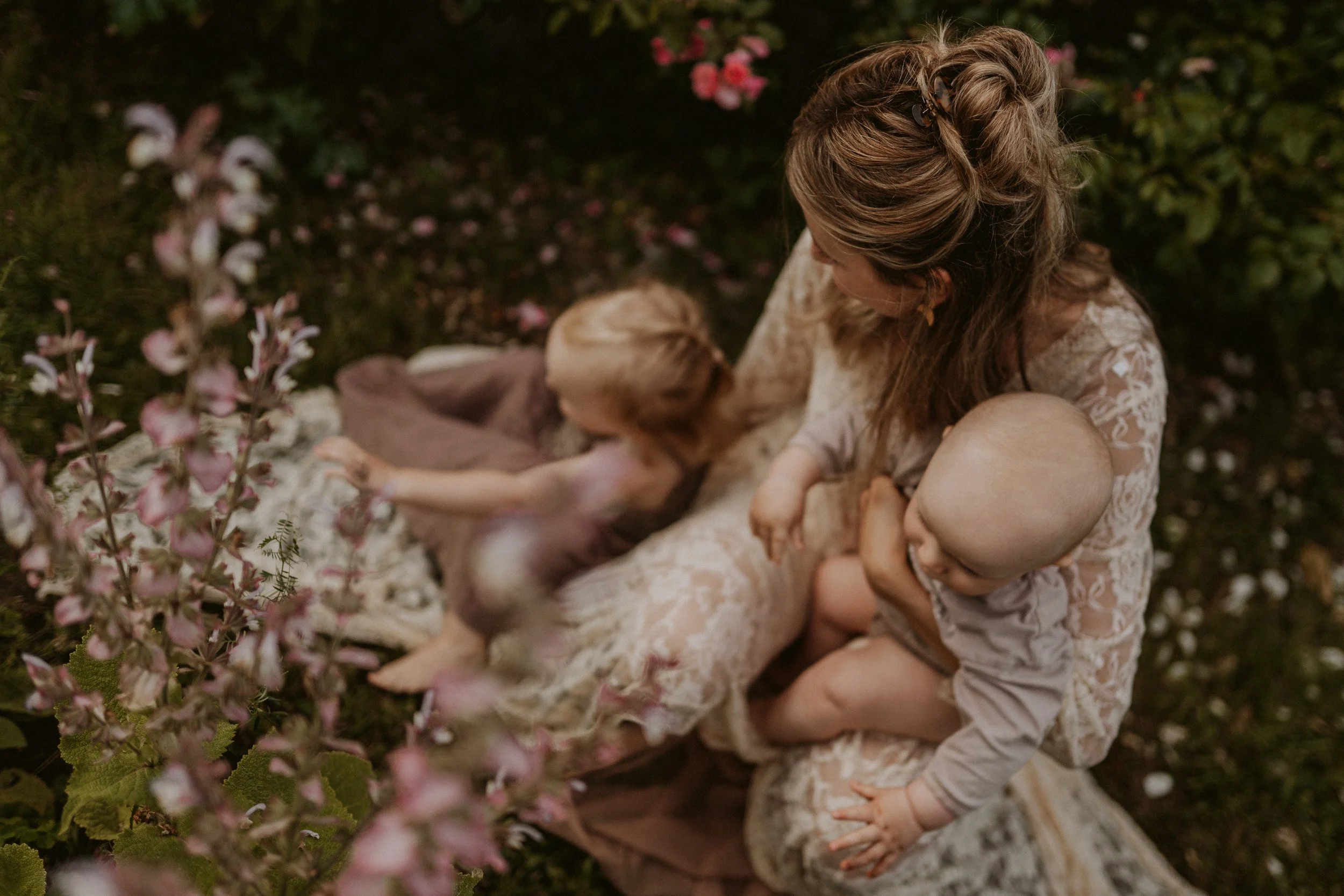 Eine Frau mit zwei kleinen Kindern sitzt im Garten, umgeben von Blumen, in einem ruhigen, intimen Moment der Nähe und Zuneigung.