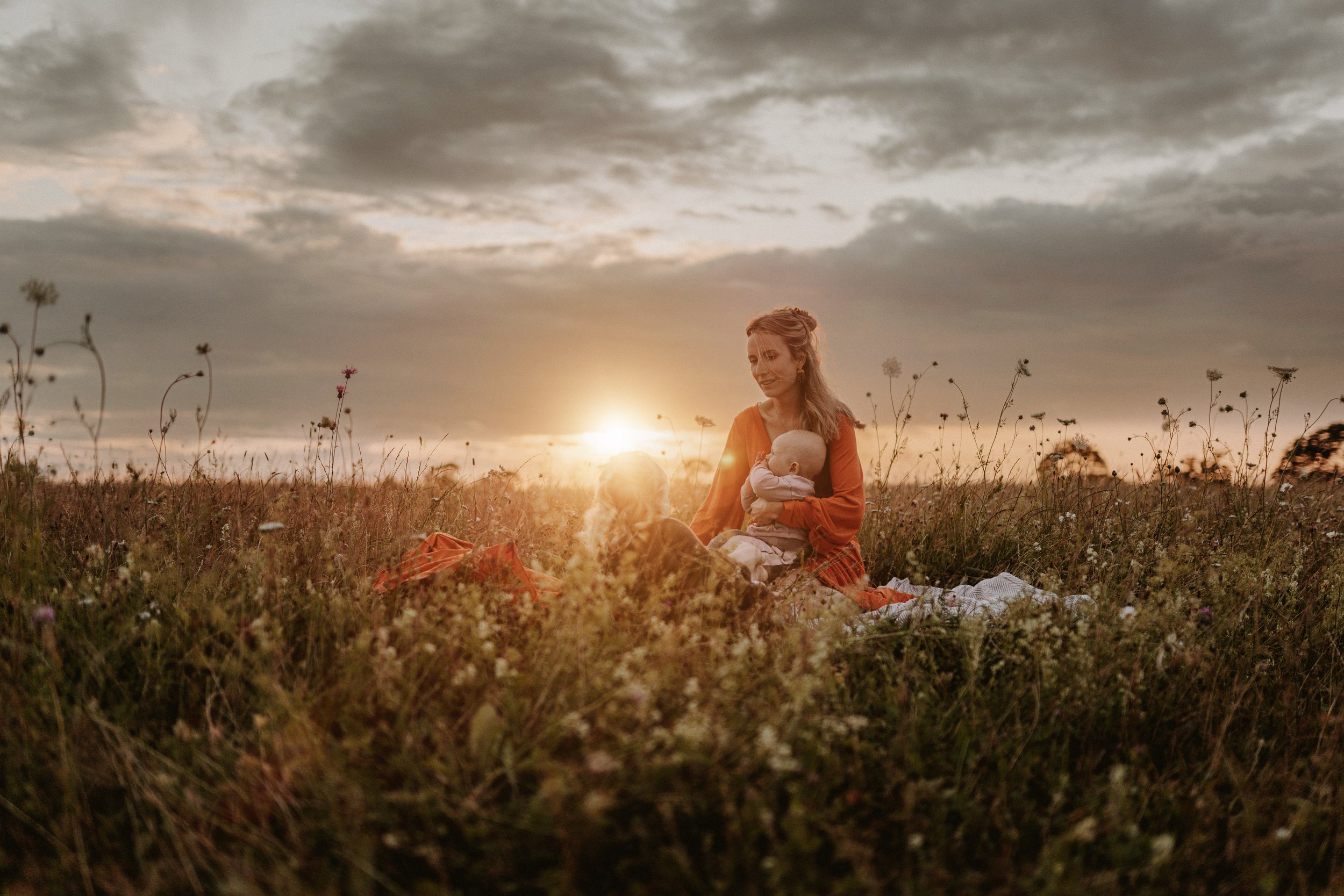 Eine Frau sitzt im Feld mit einem Baby auf ihrem Schoß bei Sonnenuntergang, umgeben von Blumen und hohen Gräsern.