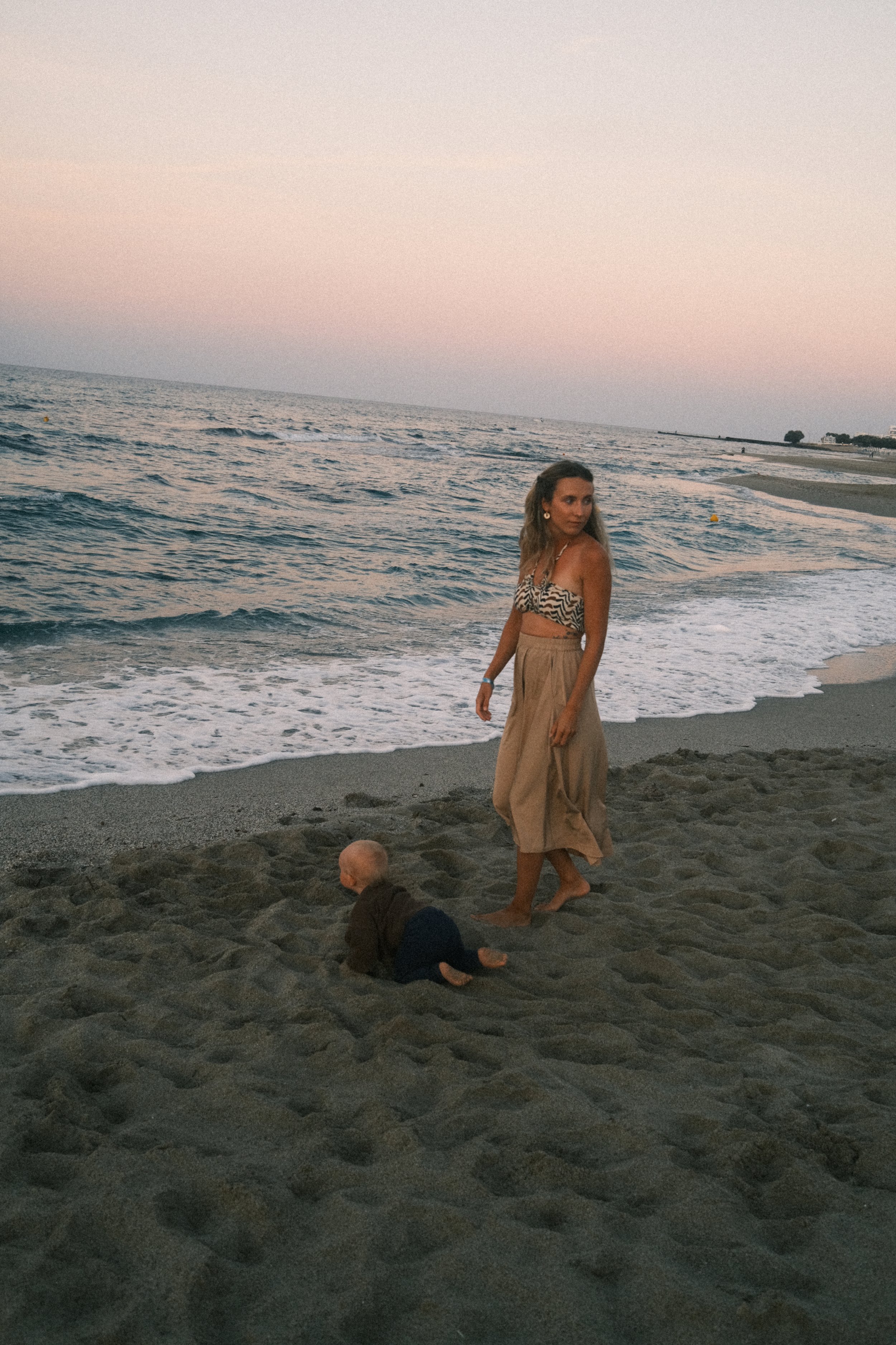 Eine Frau und ein Baby am Strand bei Sonnenuntergang. Die Frau trägt ein Tiermuster-Top und einen langen Rock, das Baby ist auf allen Vieren im Sand.