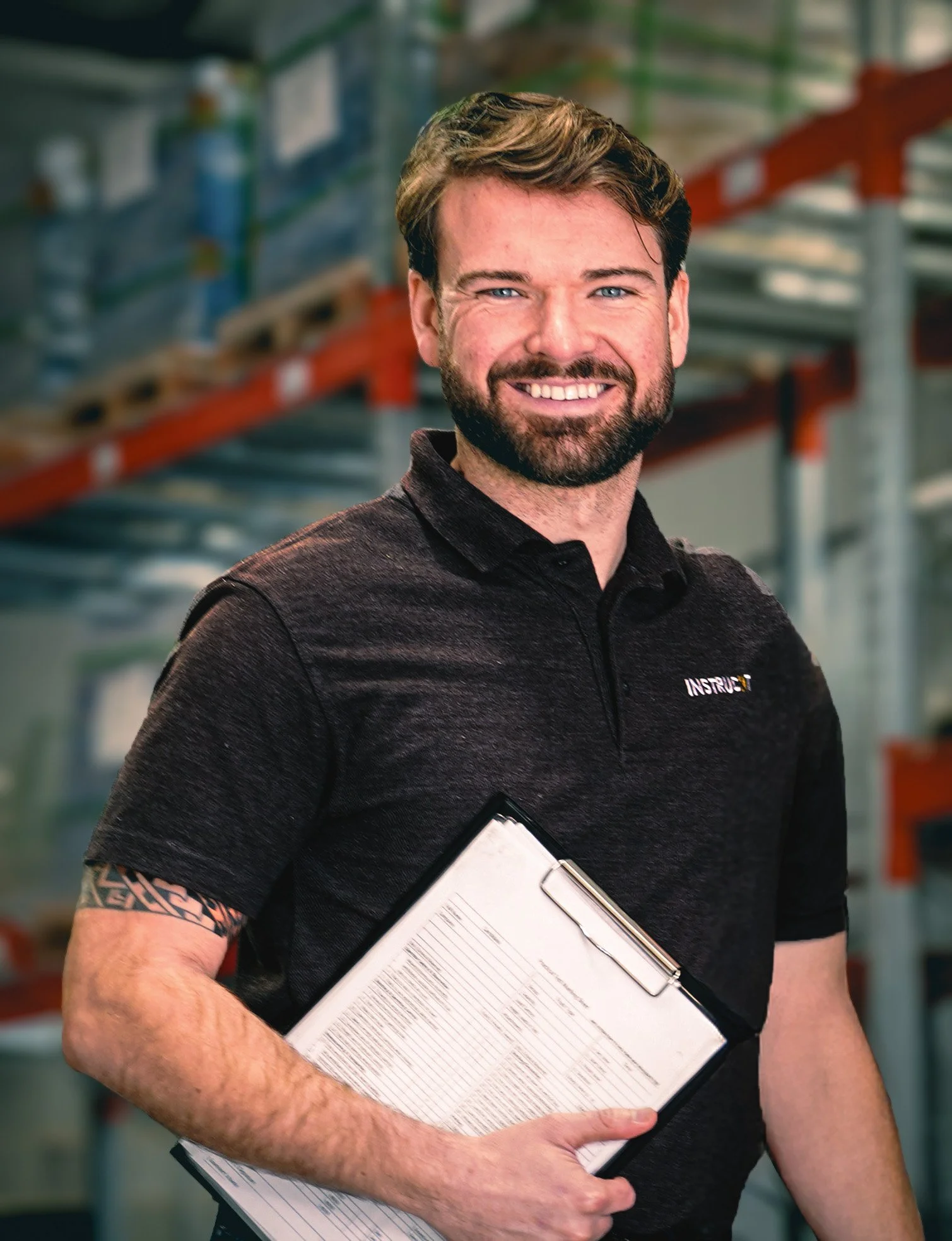 A smiling man with a beard holding a clipboard in a warehouse with metal shelves.