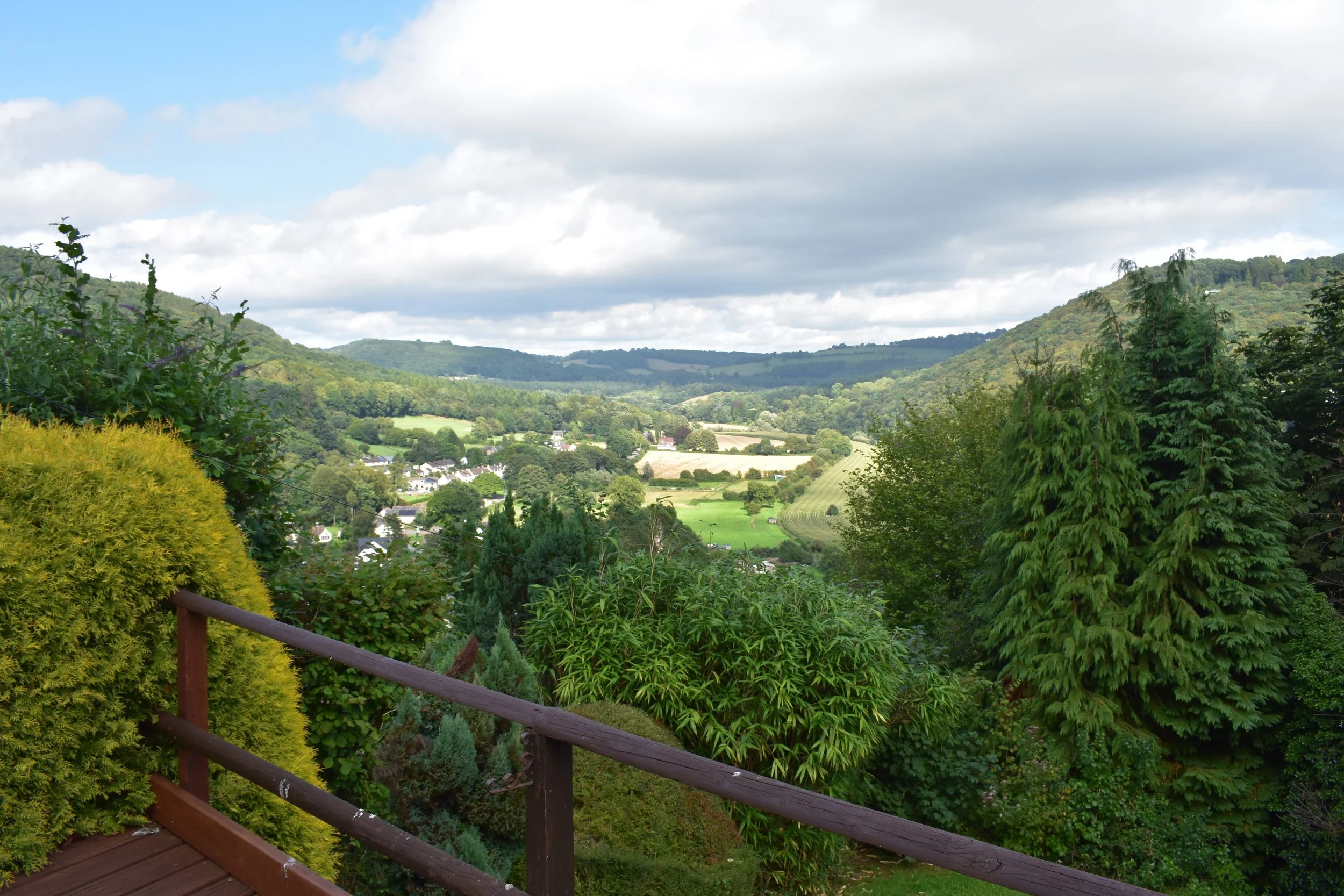 Scenic view of lush green hills, trees, and farmland beneath a partly cloudy sky, seen from a wooden balcony with a railing.