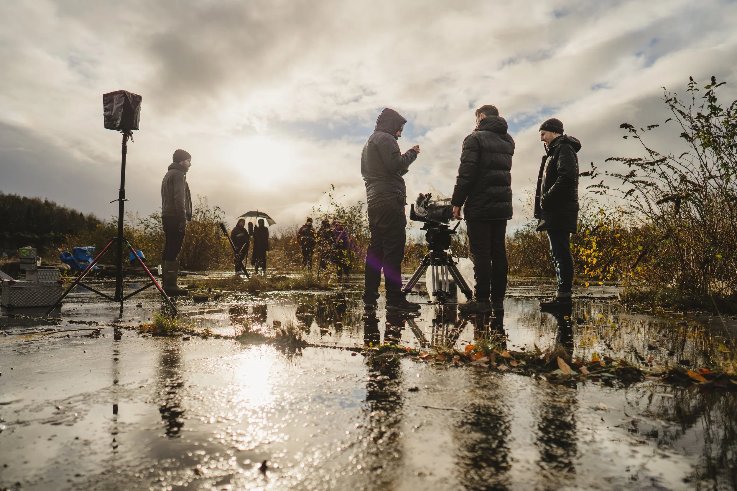 Film crew members standing in a muddy outdoor location, with a camera on a tripod, a light stand, and crew members holding equipment, during cloudy weather with sunlight breaking through.