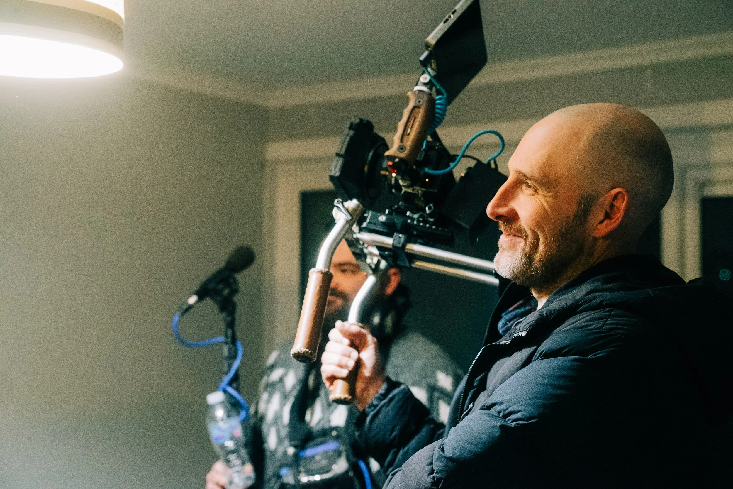 A man with a shaved head and beard holding a handheld camera rig on location in a room with overhead lighting.
