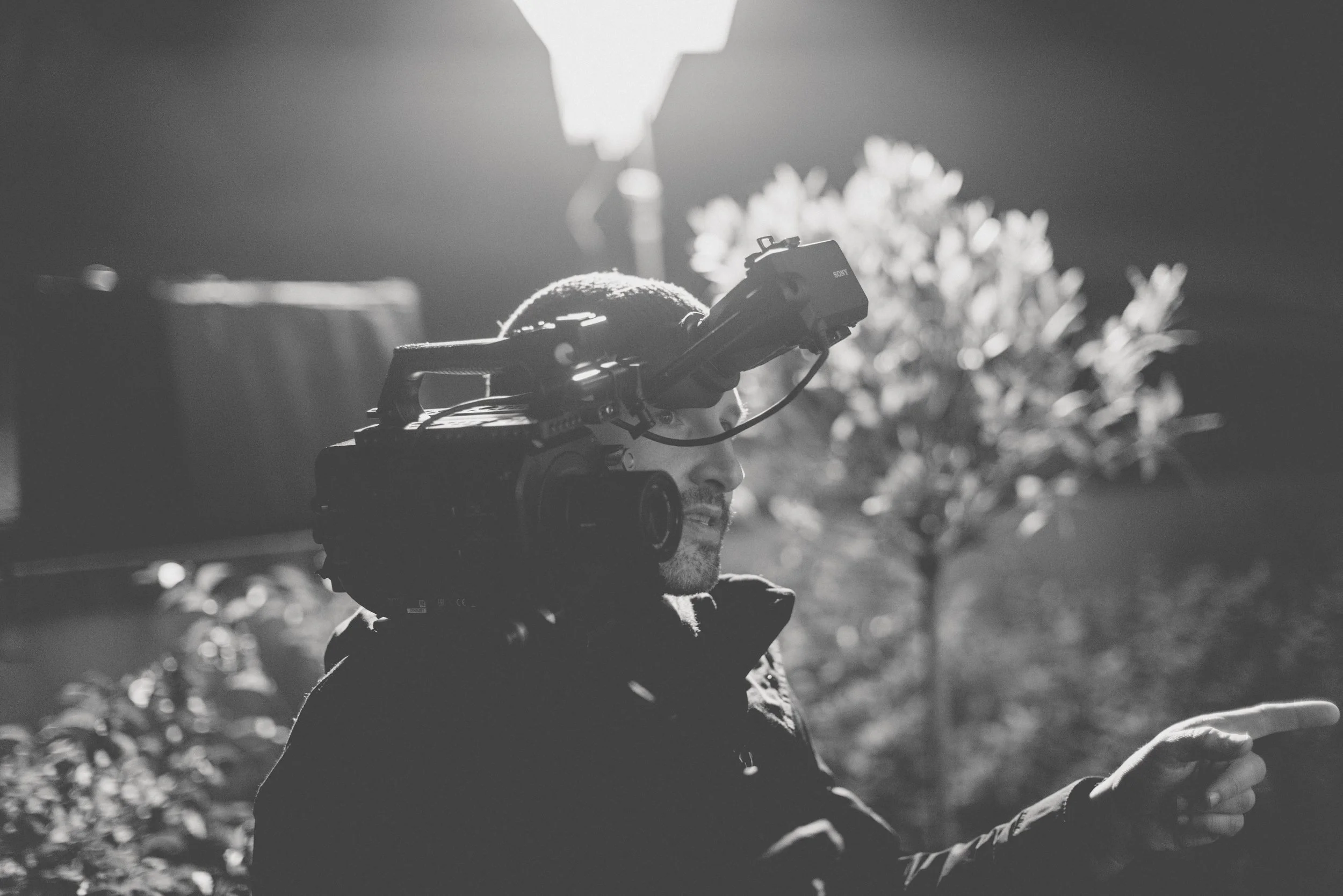 A camera man operating a video camera outdoors at night, with trees and a streetlamp in the background.
