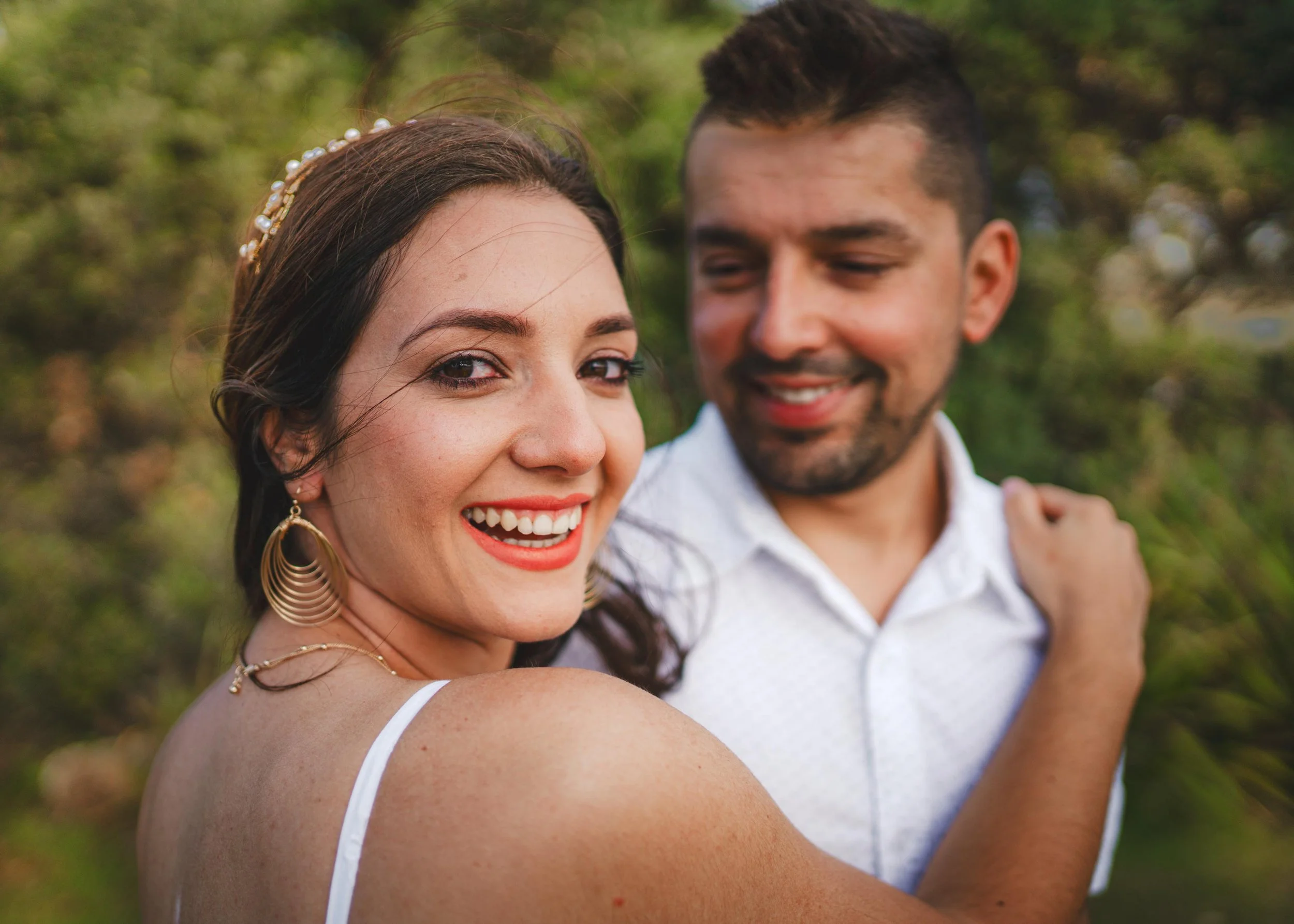 A smiling woman with dark hair, wearing gold jewelry, outdoors with a man with short dark hair and a beard, in a white shirt, on a sunny day with green blurred trees in the background.
