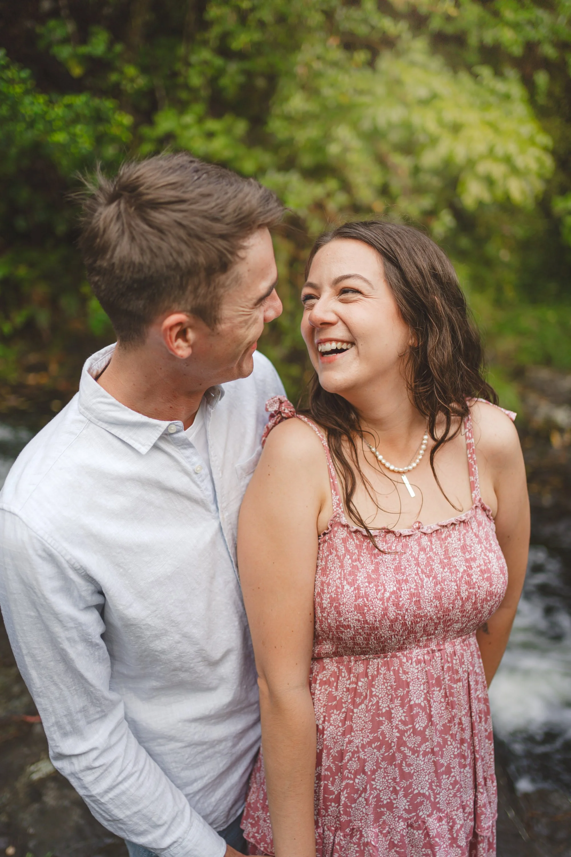 A smiling man and woman are standing close together outdoors, with a natural green background and a stream in the background.
