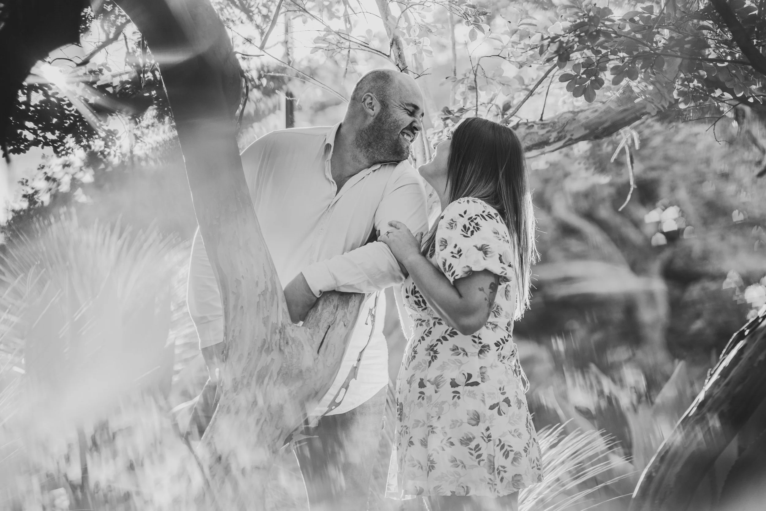 A black and white photo of a couple smiling and leaning towards each other, standing in a lush outdoor setting with trees and plants surrounding them.