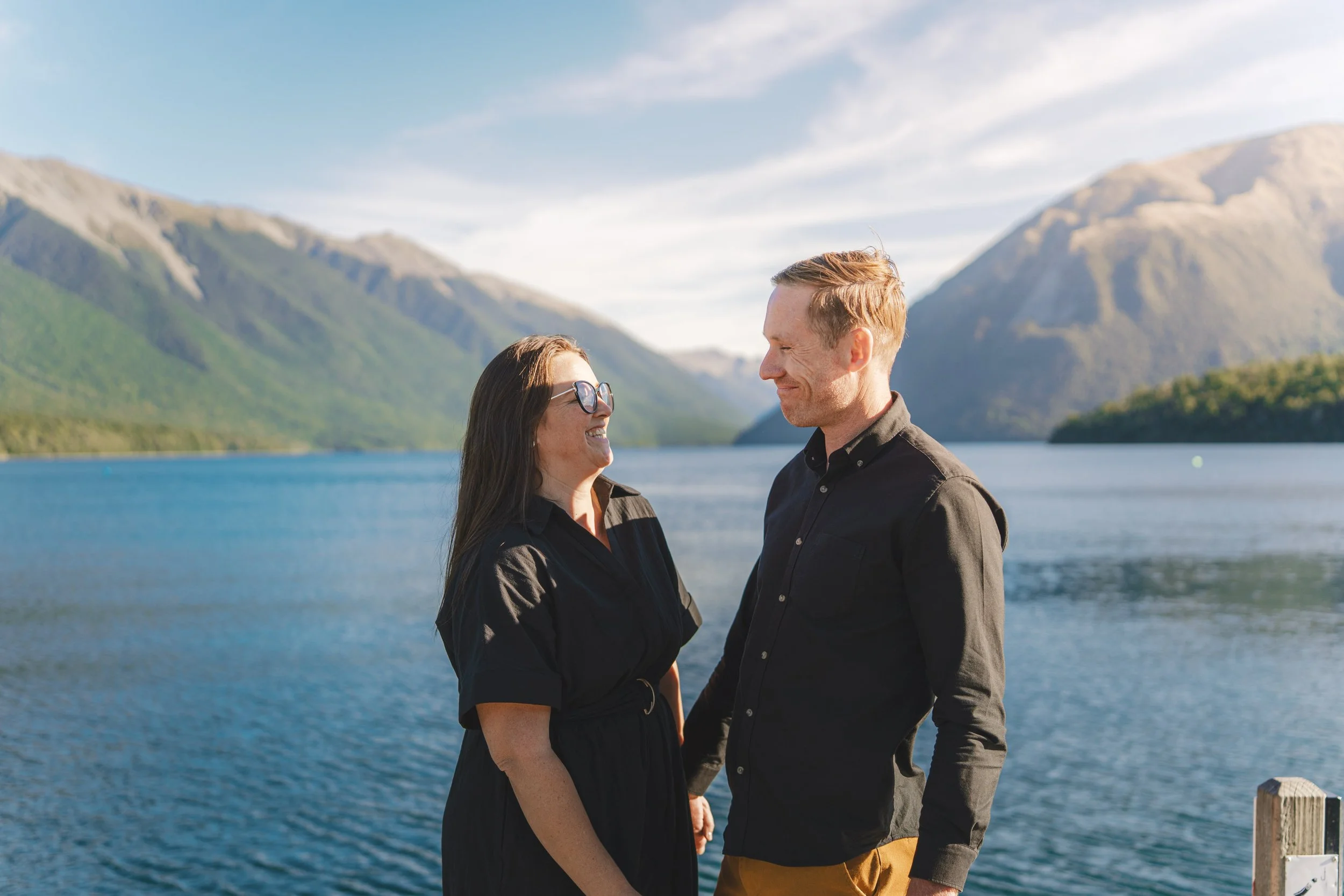 A couple holding hands and smiling at each other near a mountain lake during daytime.