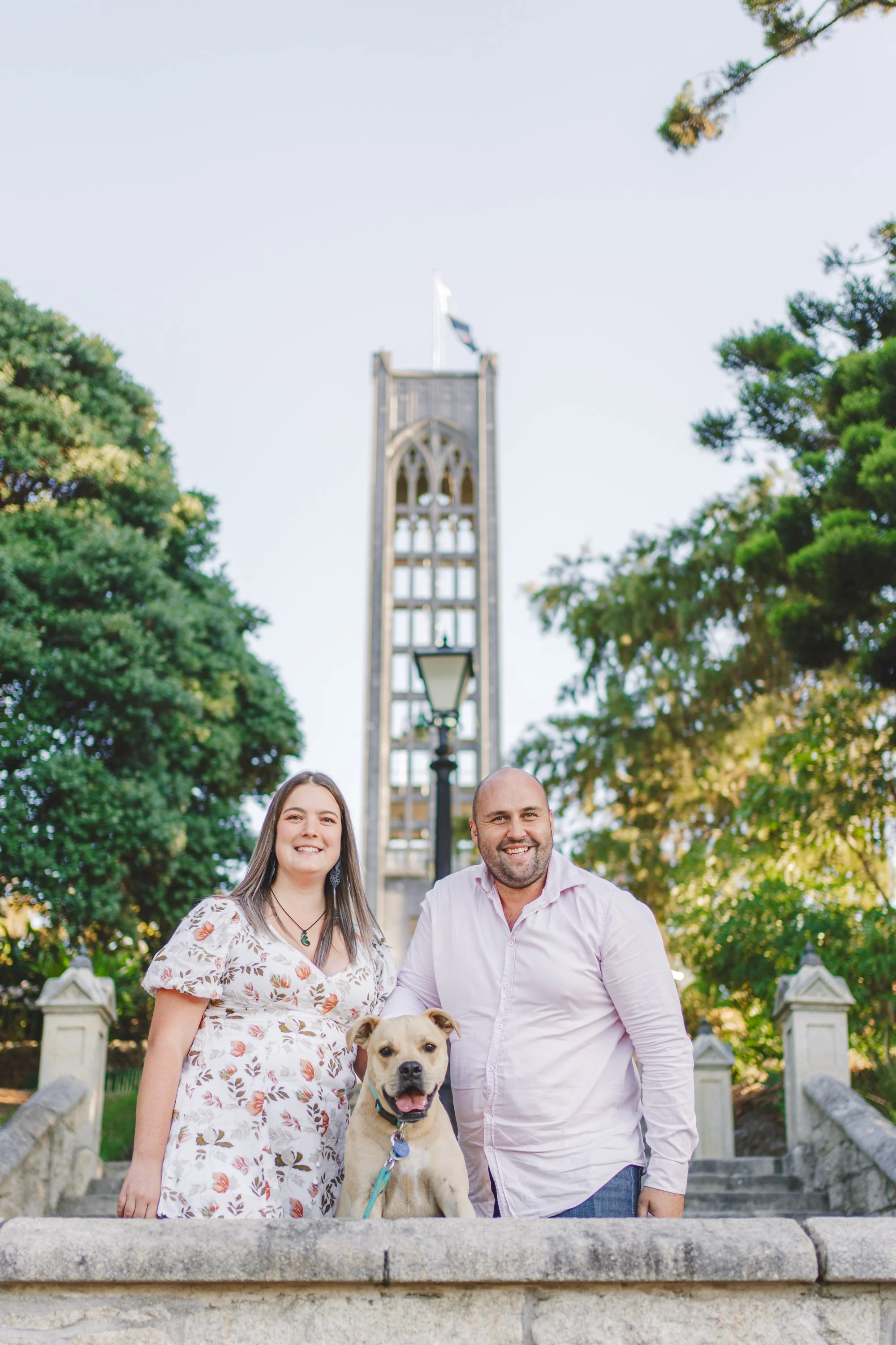 Two people, a woman and a man, standing outdoors with a dog in front of them, with a tall tower and trees in the background.