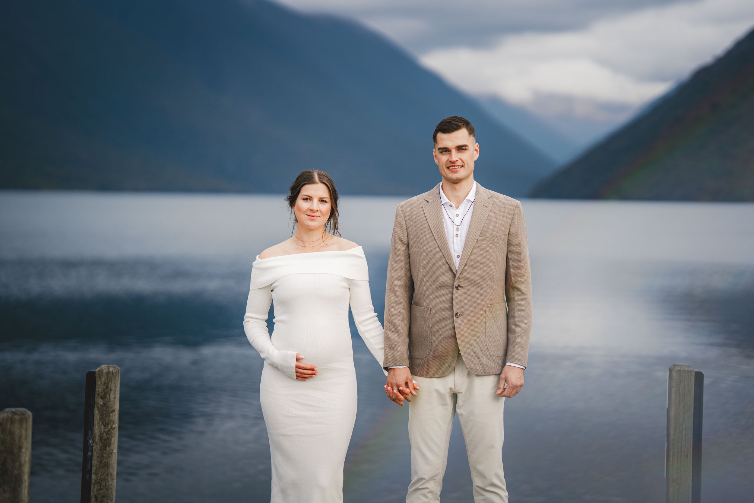 A young couple standing hand in hand by a lake with mountain scenery and cloudy sky in the background.