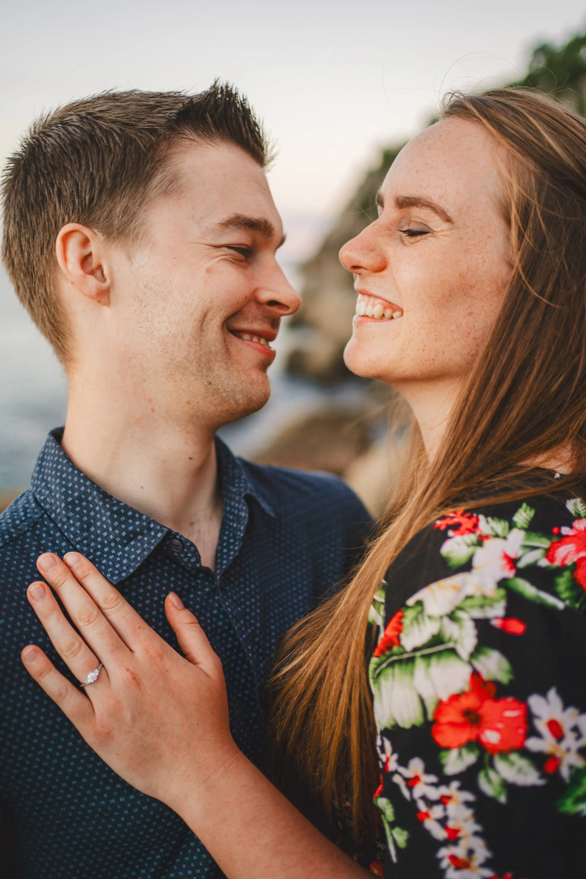 A happy couple smiling at each other, with the woman touching the man's chest, outdoors near water and rocks.