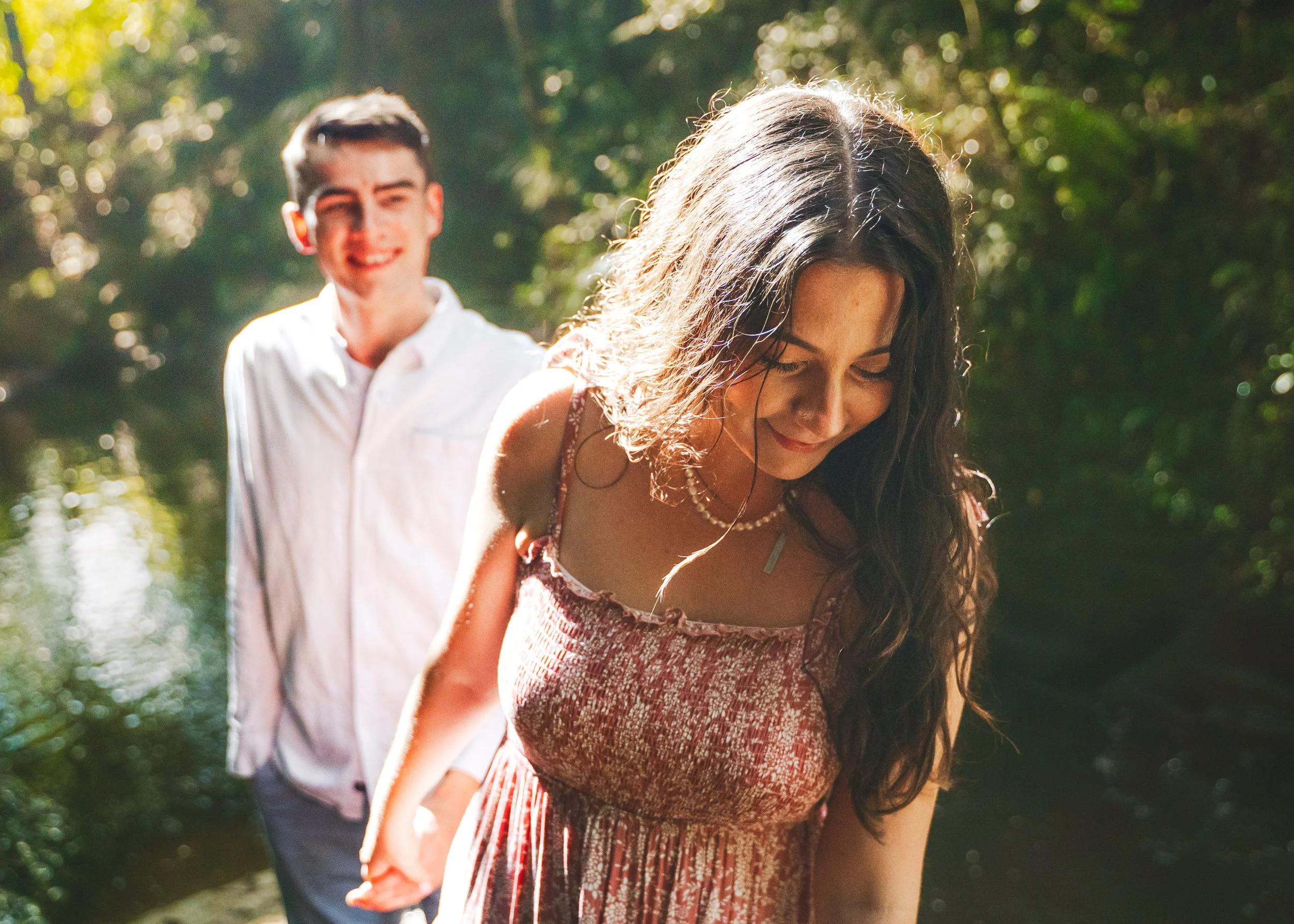 A young woman with long, curly hair wears a patterned dress and a pearl necklace, smiling and looking down. A young man in a white shirt stands behind her, smiling with a blurred background of green foliage and sunlight.