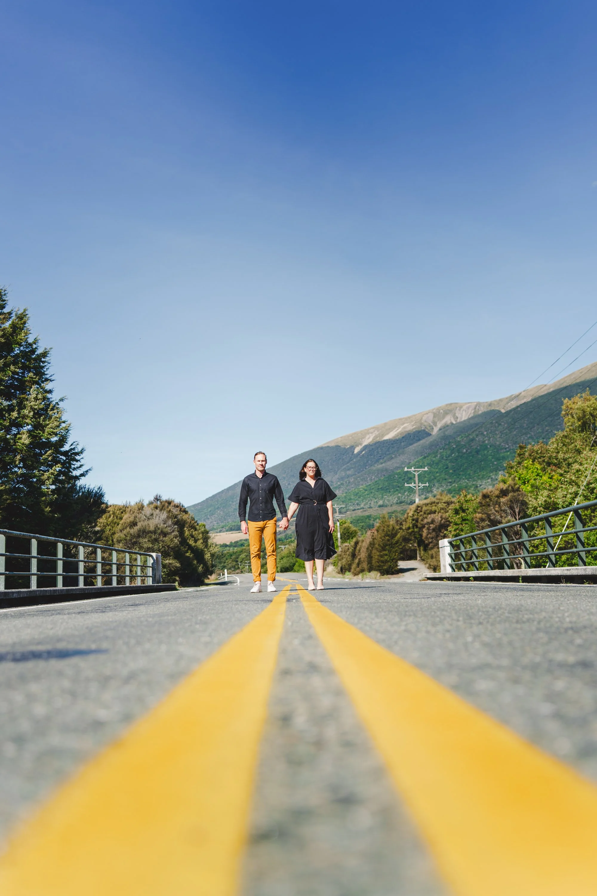 Man and woman holding hands walking on a rural road between green trees and mountains in the background during daytime.