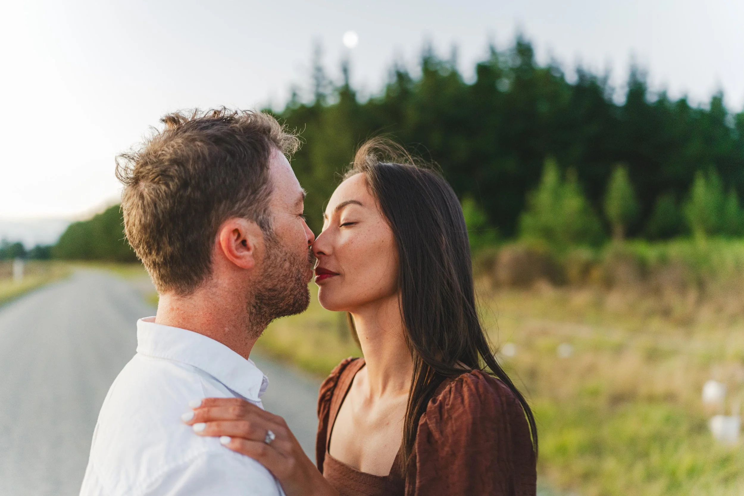 A man and woman about to kiss outdoors on a road with trees in the background