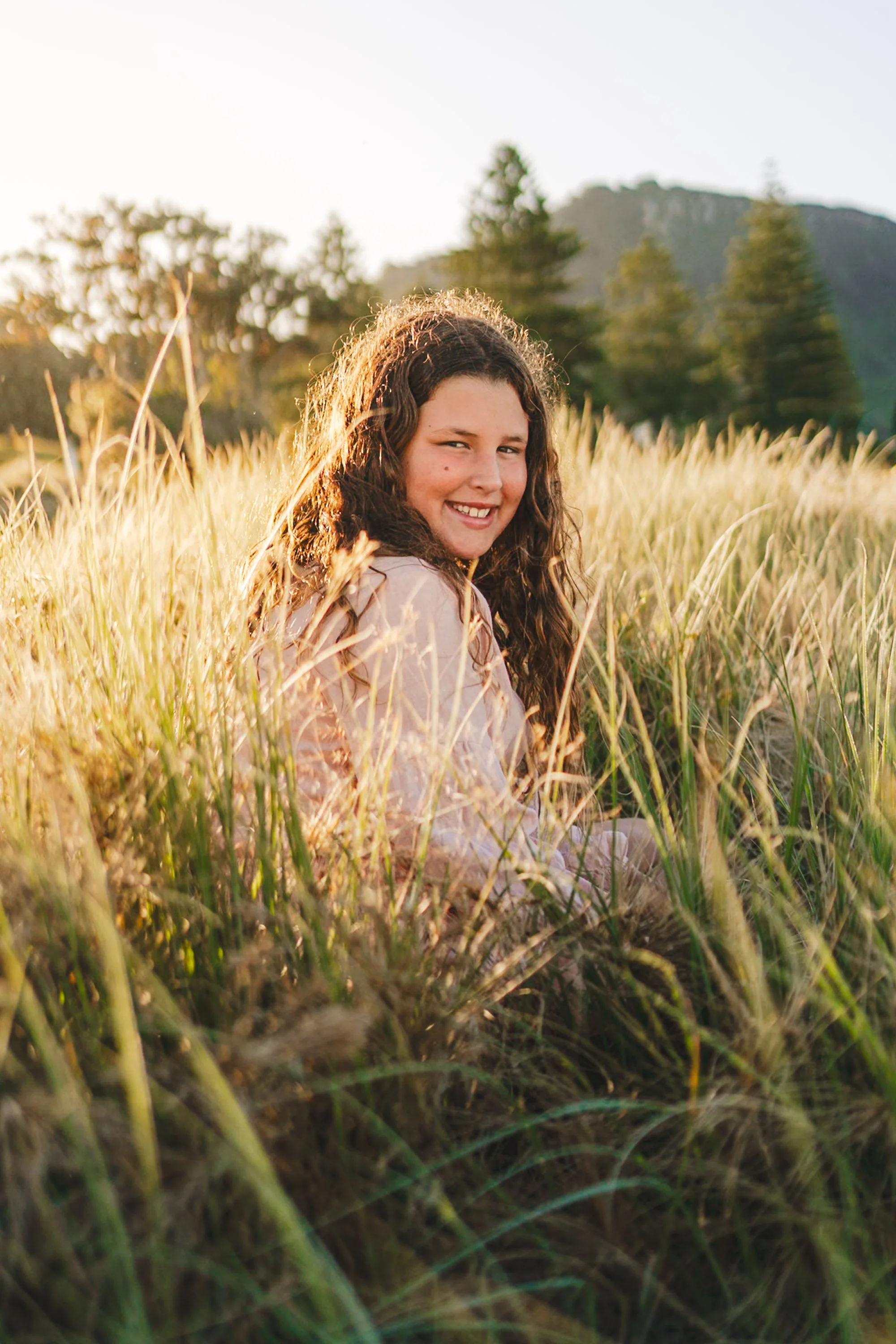 A young girl with long, curly hair smiling and sitting in a field of tall grass at sunset.