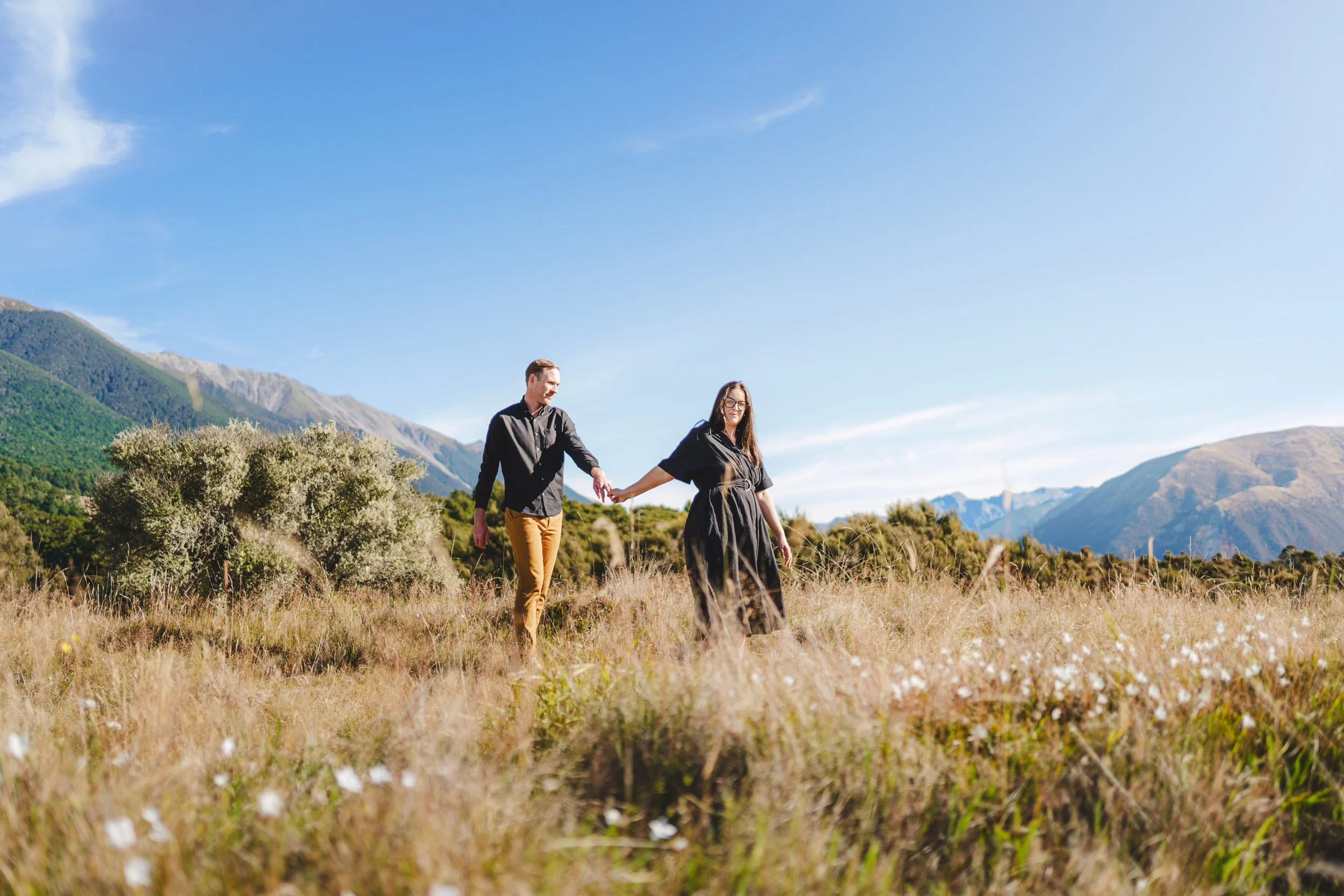 A couple walking hand-in-hand through a grassy field with mountains in the background and a clear blue sky.