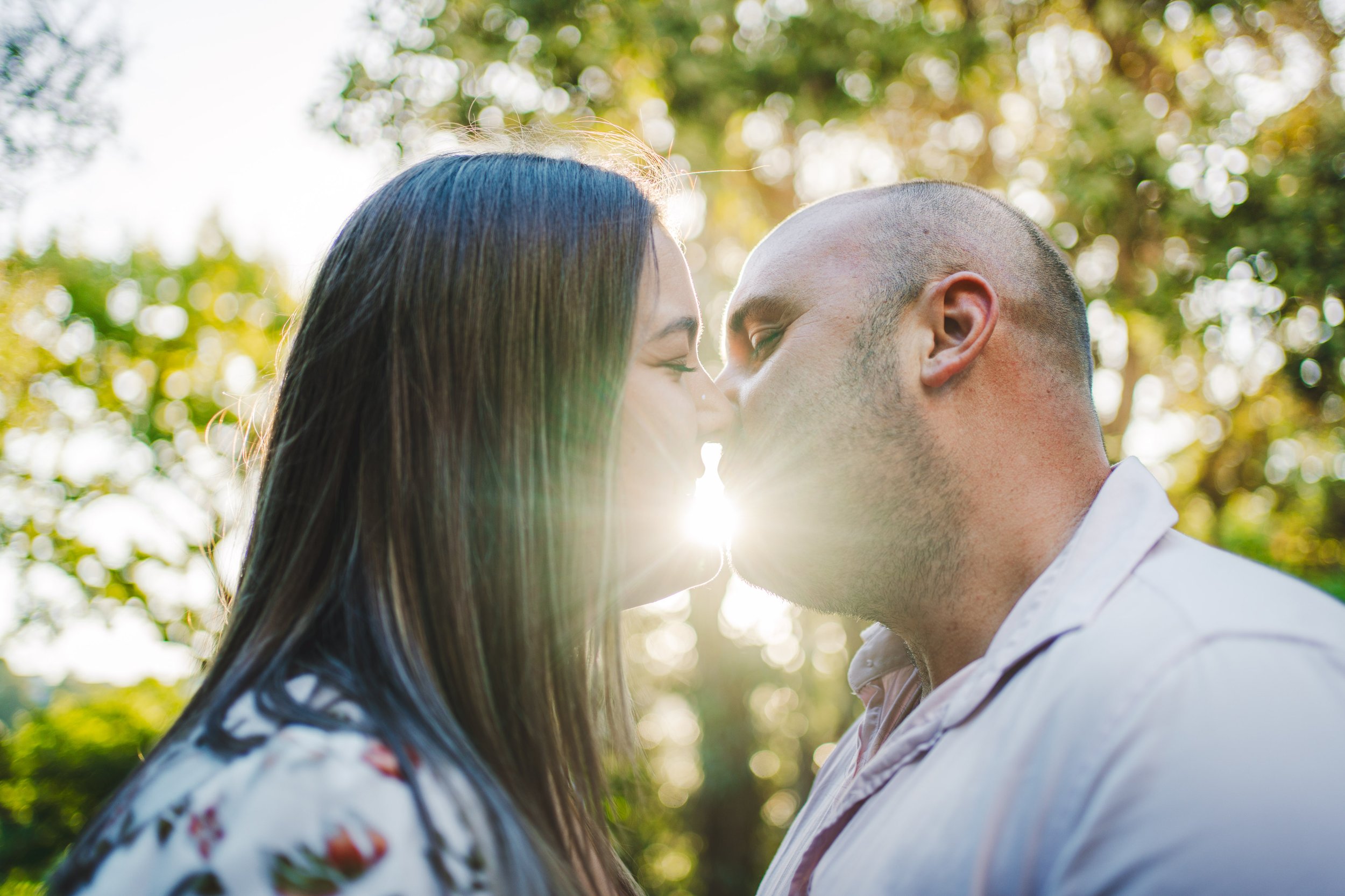 A couple is about to kiss outdoors with sunlight shining behind them and trees in the background.
