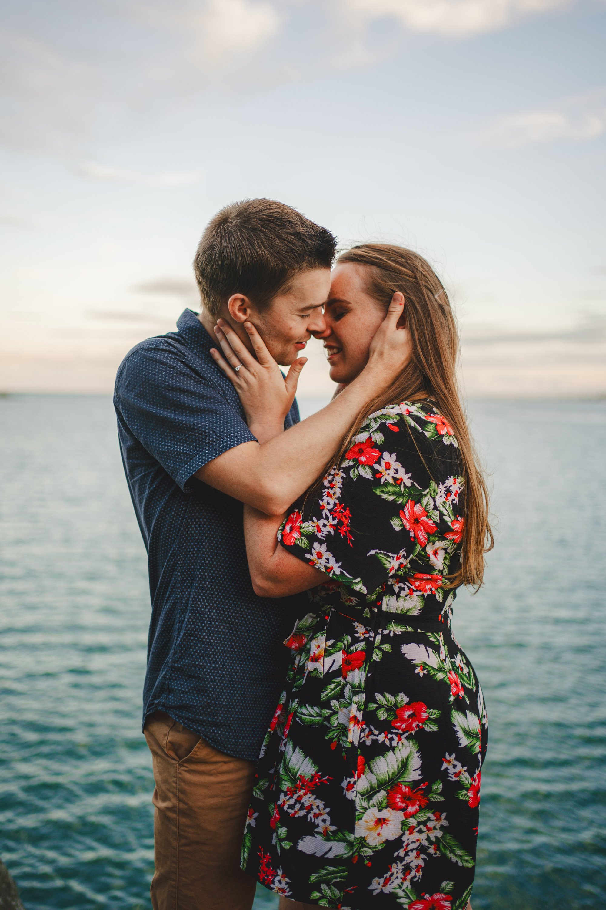 A couple embraces closely near a body of water at sunset, with sky and clouds in the background.